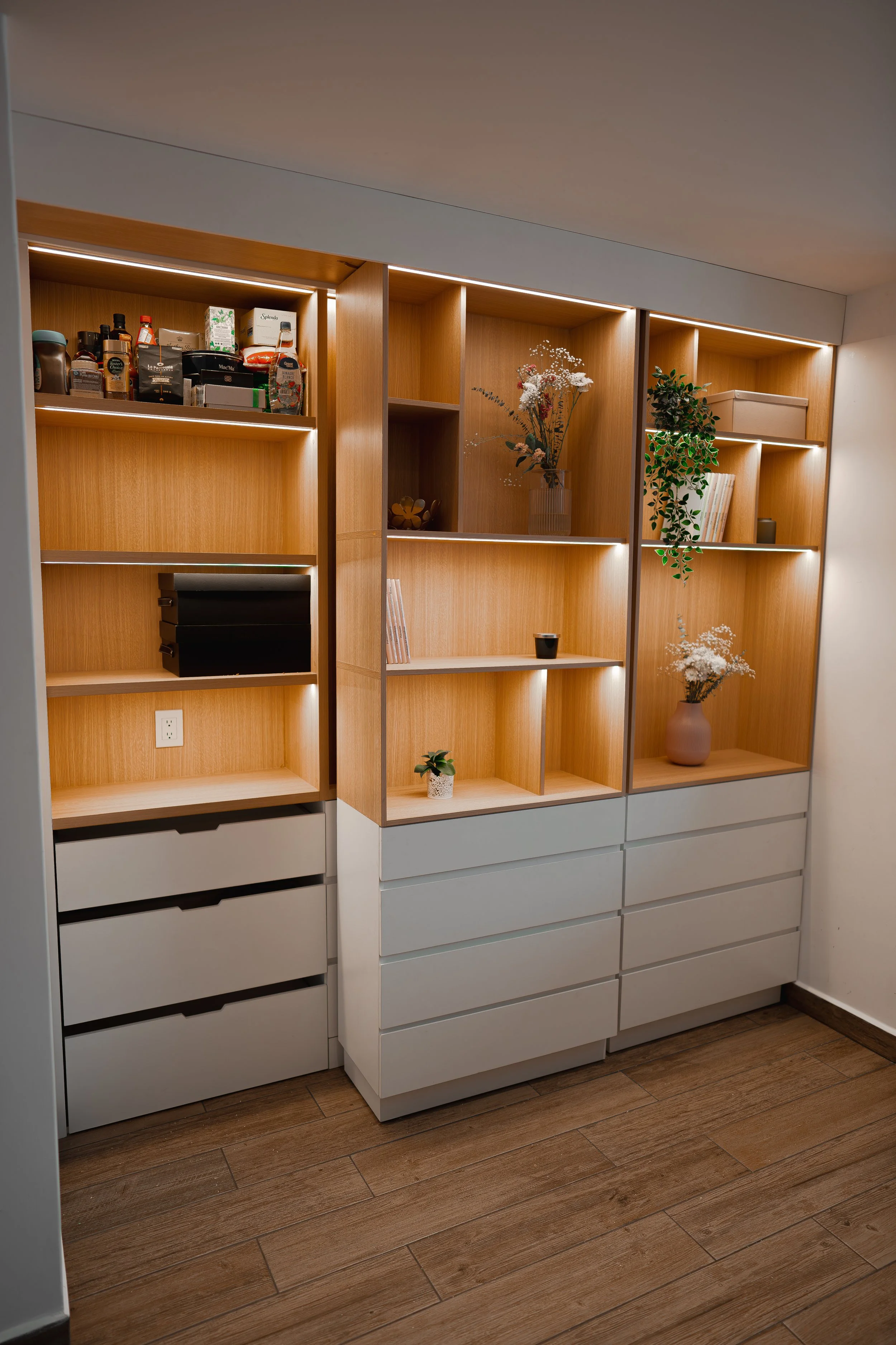 Estantes de madera con objetos decorativos, plantas y caja, junto a un piso de madera y pared blanca.