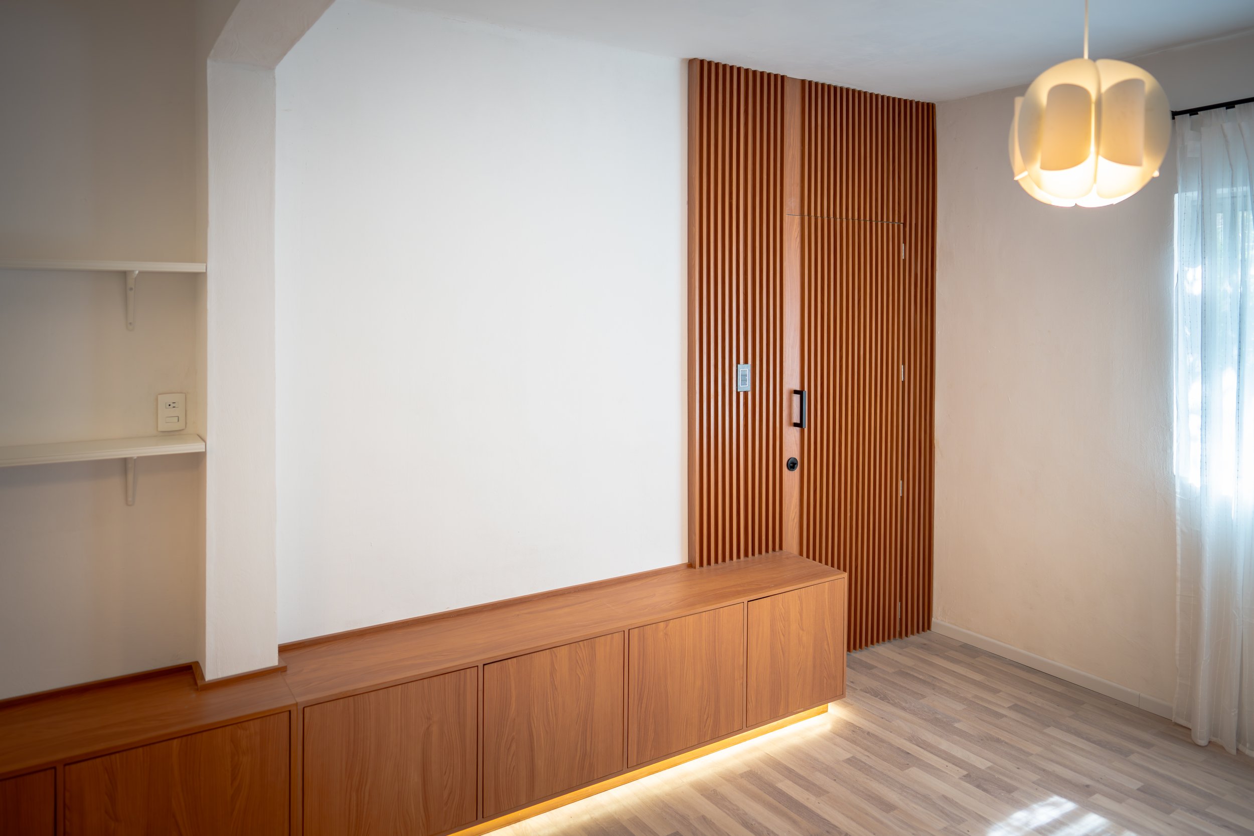 Entrada de una sala con pared blanca, mueble de madera en la parte inferior, con paneles de madera vertical en la pared y una lámpara decorativa de forma redonda suspendida del techo. La luz de la lámpara ilumina sutilmente la habitación, junto a cortinas blancas que dejan entrar luz natural.