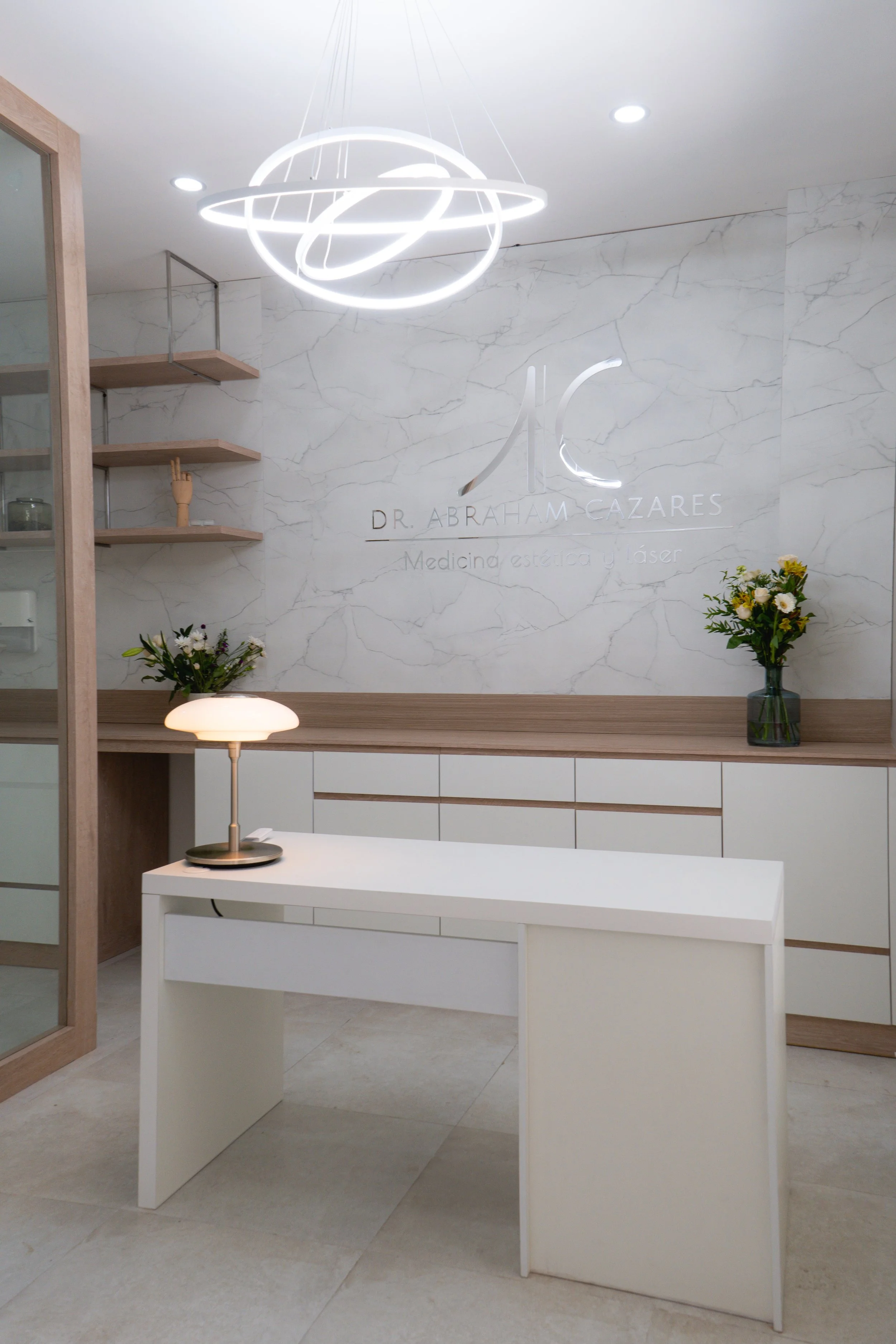 Recepción moderna en clínica estética con nombre 'Dr. Abraham Cazares', decorada con plantas y lámpara de mesa, paredes de marmol y estanterías en tonos madera, con un diseño limpio y minimalista.