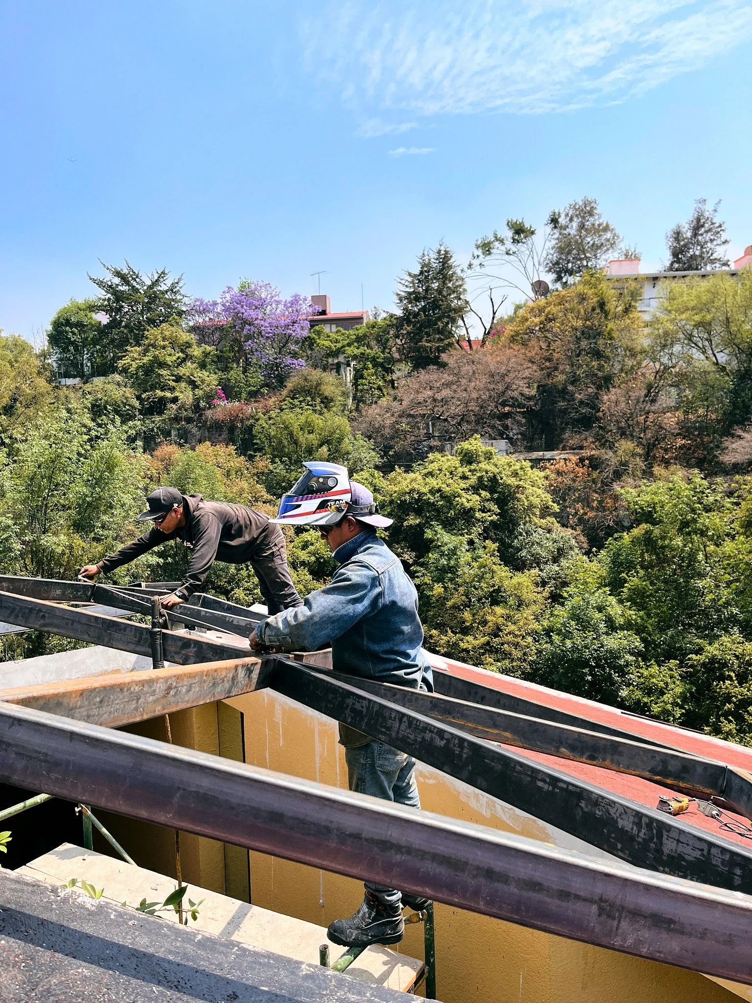 Dos trabajadores en un andamio trabajando en una estructura de construcción, con árboles y casas en el fondo bajo un cielo despejado.