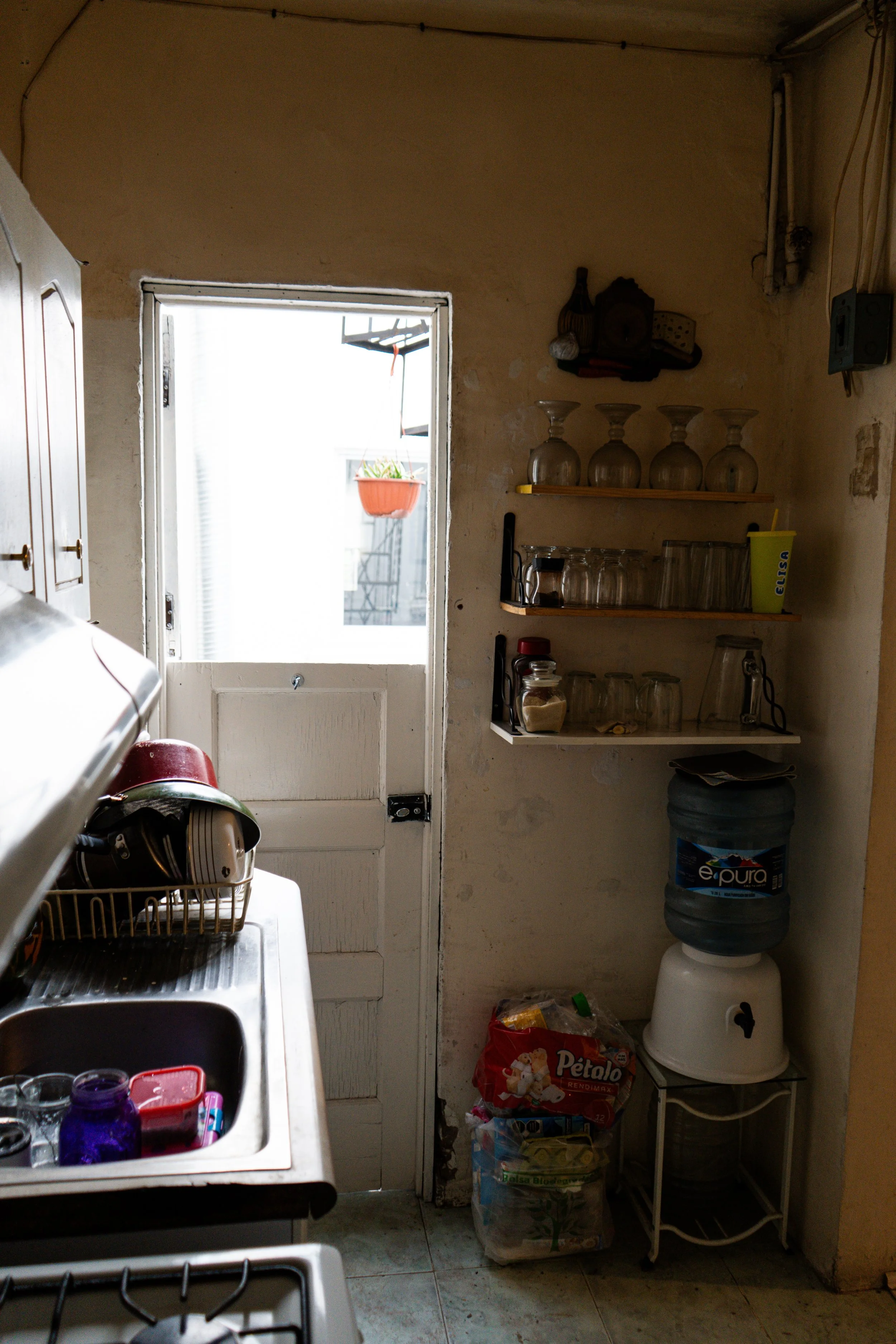 Interior de una cocina con estantes, utensilios, una llave de agua, una olla de agua potable, paquetes de alimentos y una puerta que da a un balcón con una maceta colgante con plantas.