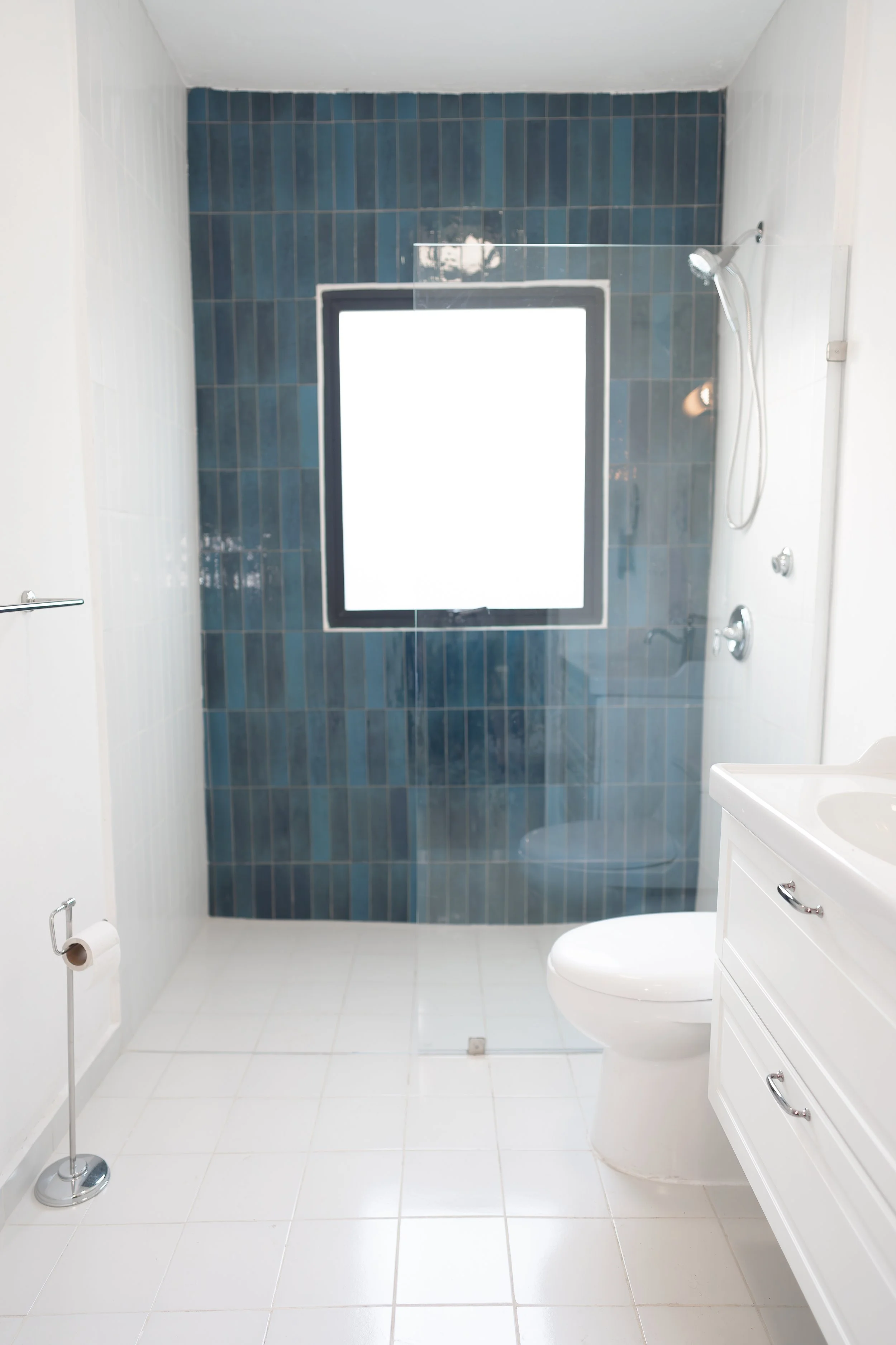 Baño con ducha, azulejos azules en la pared de la ducha, ventana con cortina blanca, inodoro, armario con tres cajones, lavabo blanco, papel higiénico, azulejos blancos en el suelo.