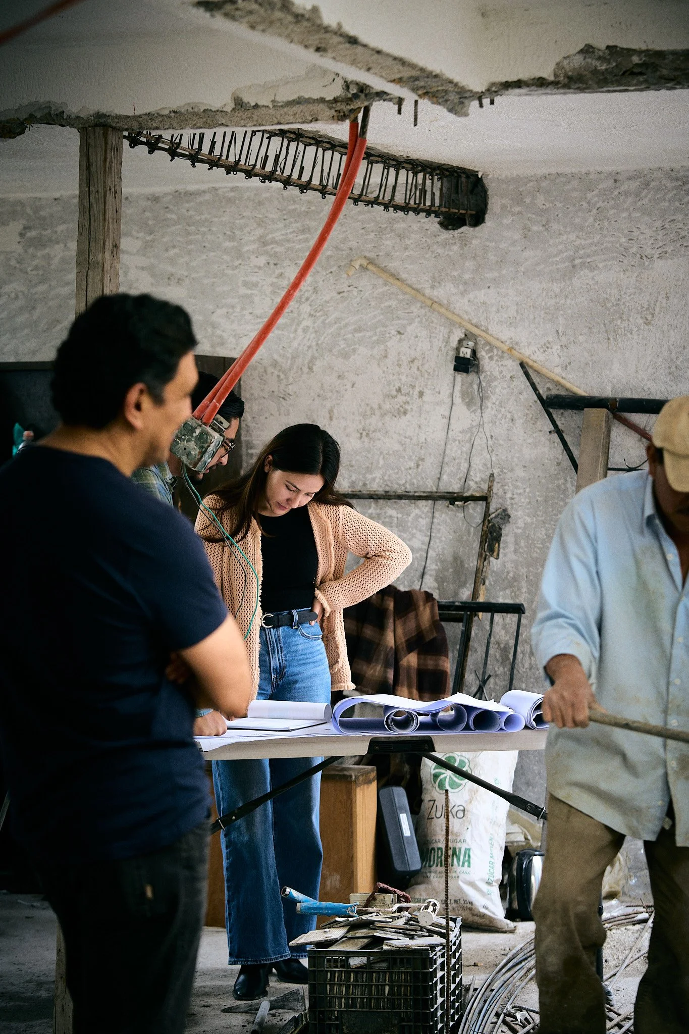 Grupo de personas en una construcción inspeccionando planos y materiales, con paredes sin terminar y herramientas alrededor.