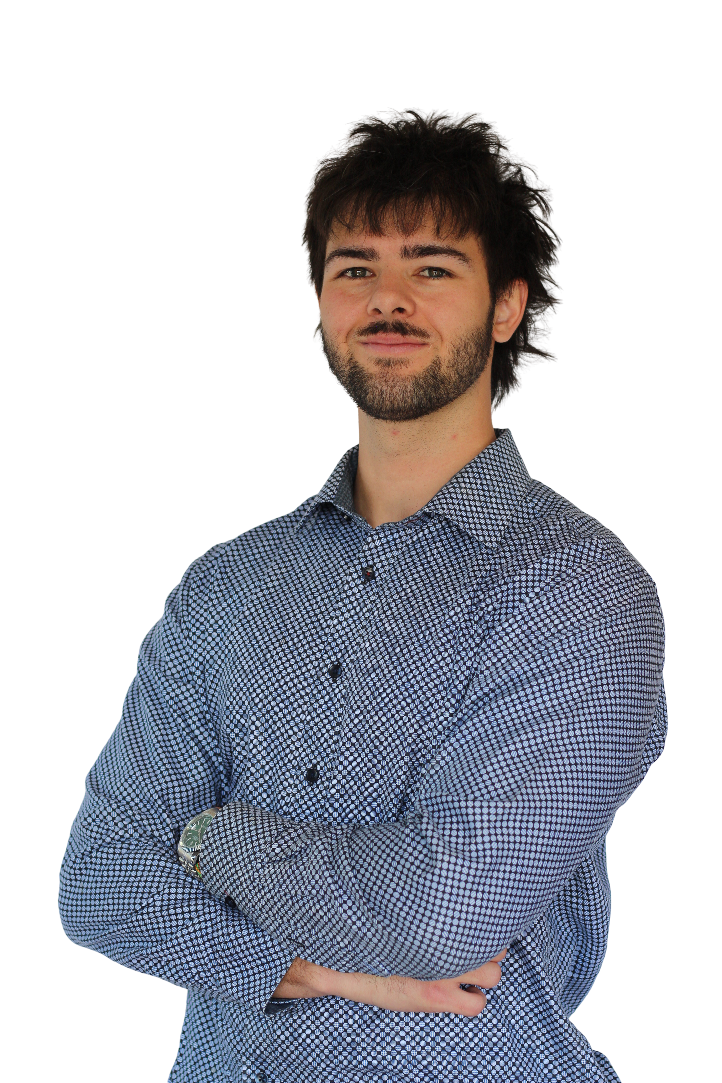 A young man with dark, curly hair, a beard, and wearing a blue patterned button-up shirt standing with arms crossed, smiling slightly.