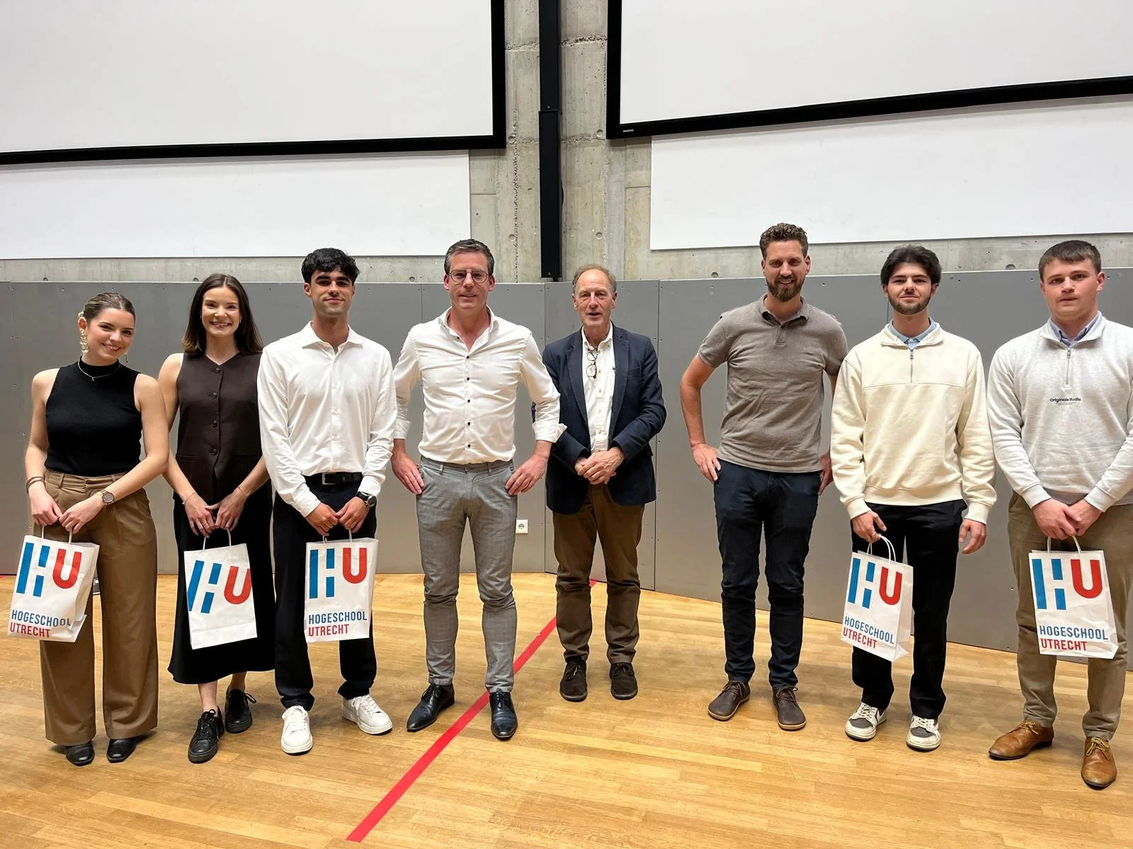 Group of nine people standing in a gymnasium, holding bags that say 'Hogeschool Utrecht'. They are dressed semi-formally, with some wearing casual clothing. The setting appears to be an academic or professional event.