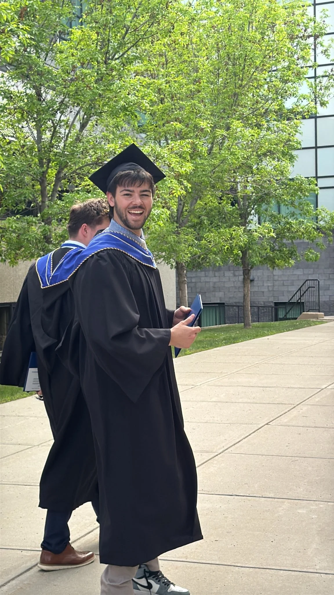 A young man in a graduation gown and cap smiling and holding a diploma, walking outdoors on a sunny day with trees and a building in the background.
