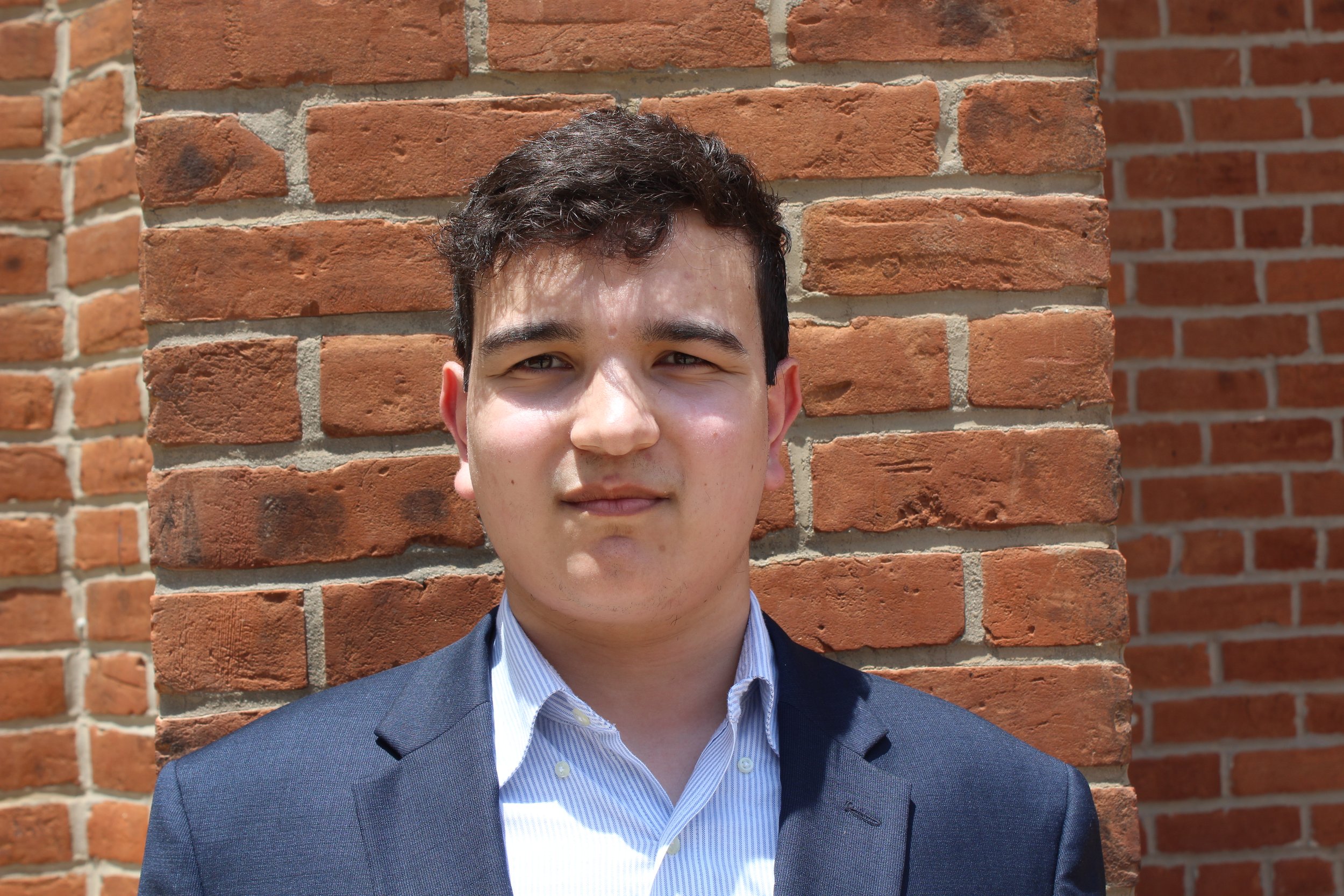 A young man in a suit smiling outdoors in front of a brick wall.