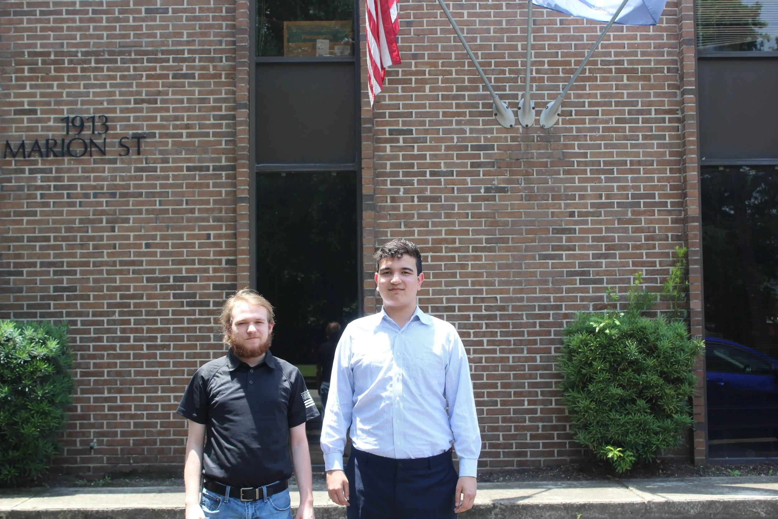 Two young men dressed in formal attire standing outdoors in front of a brick building with the address 1913 Marion Street visible. An American flag and a blue and white flag are mounted on the building. There are bushes on either side of the young men.