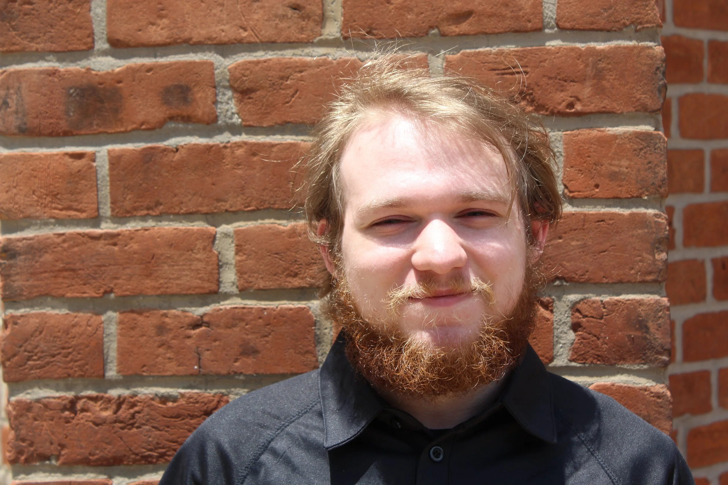 Close-up of a young man with light skin, curly blond hair, and a beard, smiling slightly, standing against a brick wall on a sunny day.