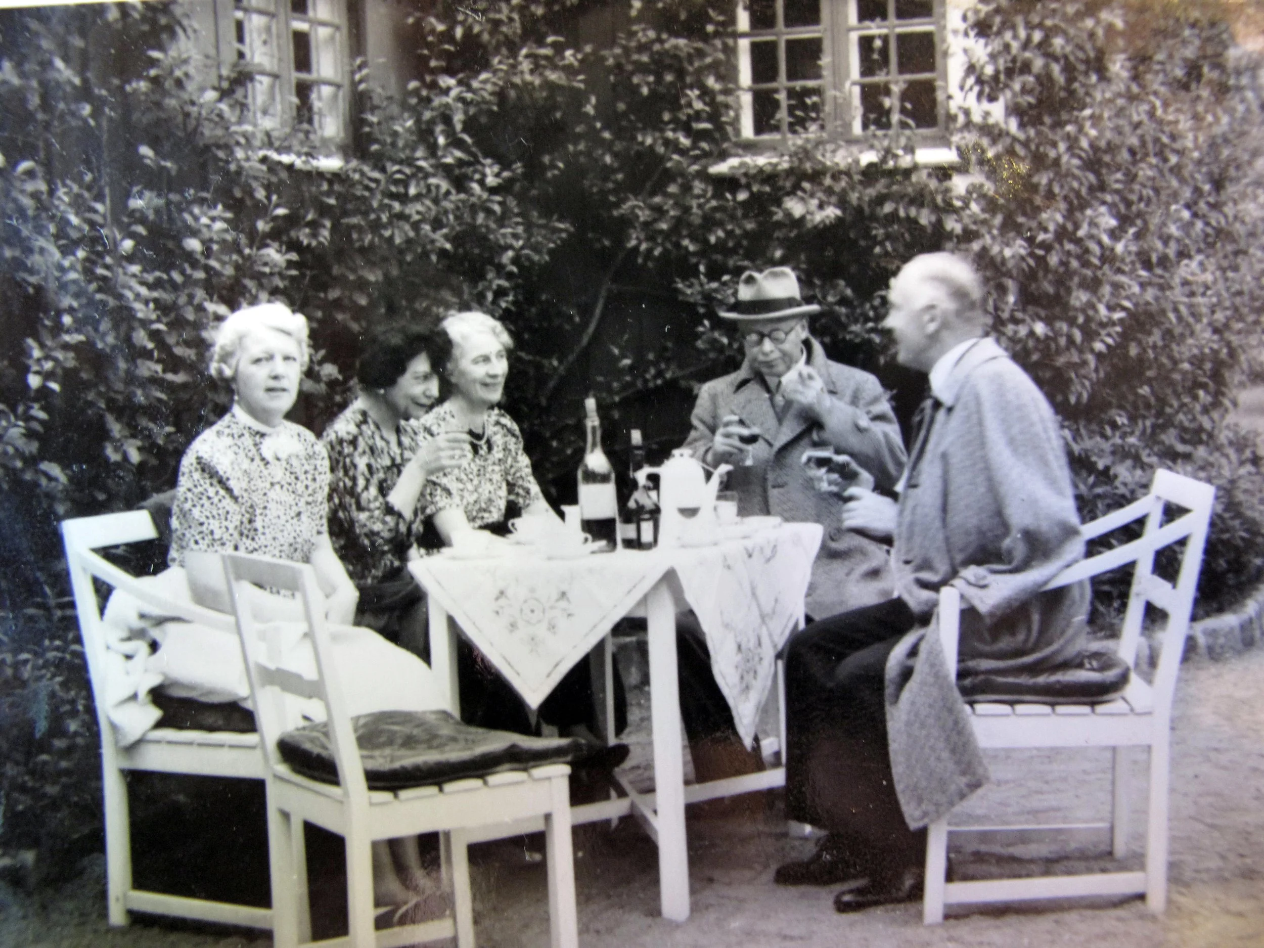 Svartvit fotografi av fem personer som äter och dricker vid ett utomhusbord, med stora fönster och buskar i bakgrunden.