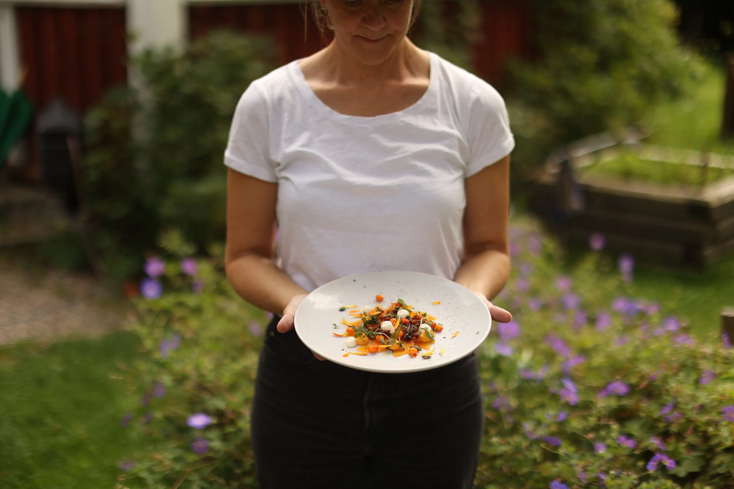 Kvinna i vit T-shirt håller en tallrik med en grönsaksrätt utomhus i en trädgård.