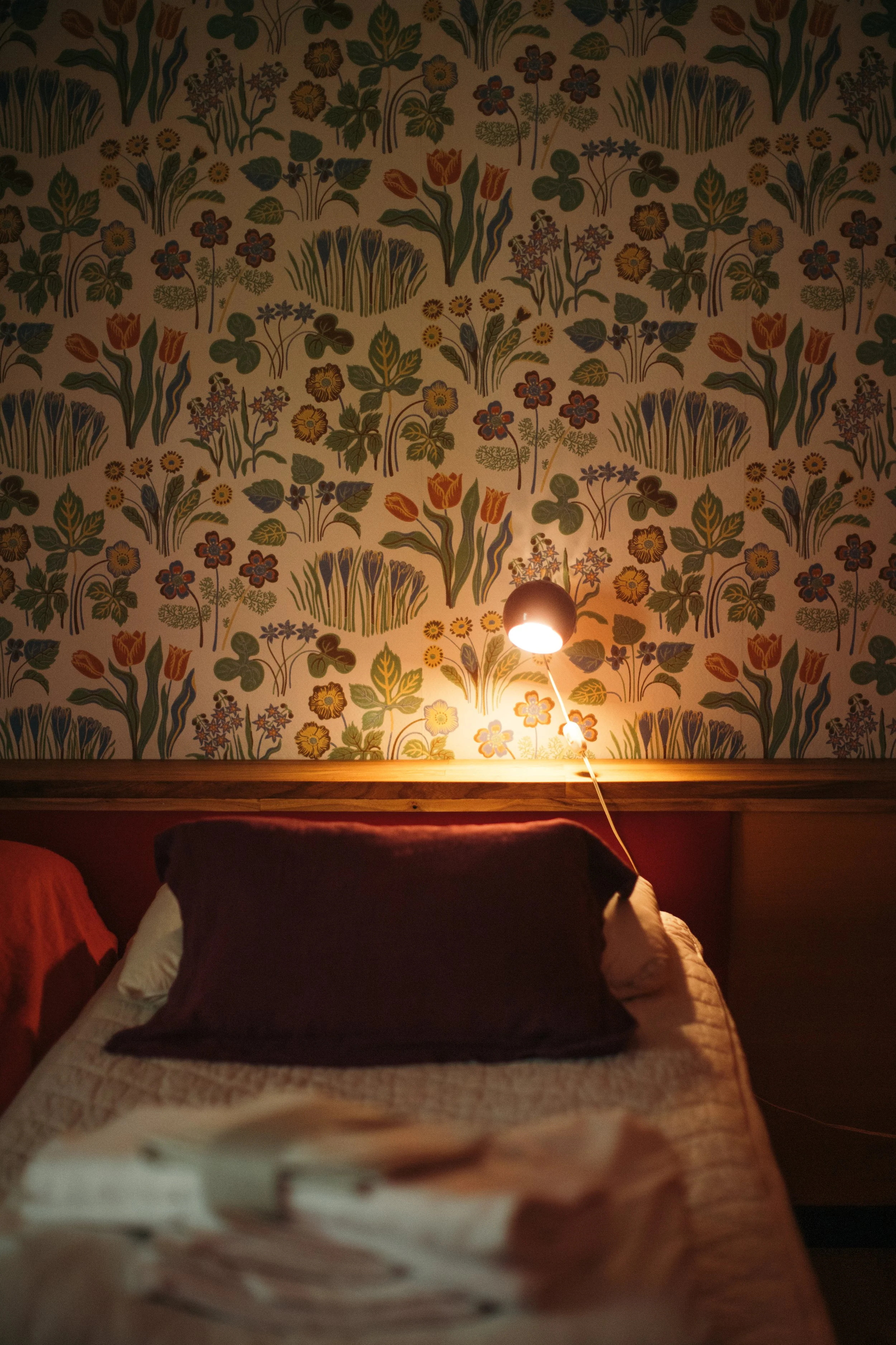 Säng med mörkblå och beige kuddar framför en vägg med blommönster. En vägglampa lyser varm ljus på lakanen.