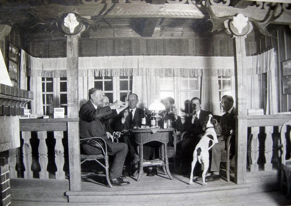 Fem män i kostym sitter runt ett bord inomhus på en gammal bild, några av dem röker, och en hund står på sina bakben vid ett av sängarna på en av männen. Det finns en lampa på bordet och flera fönster i bakgrunden.
