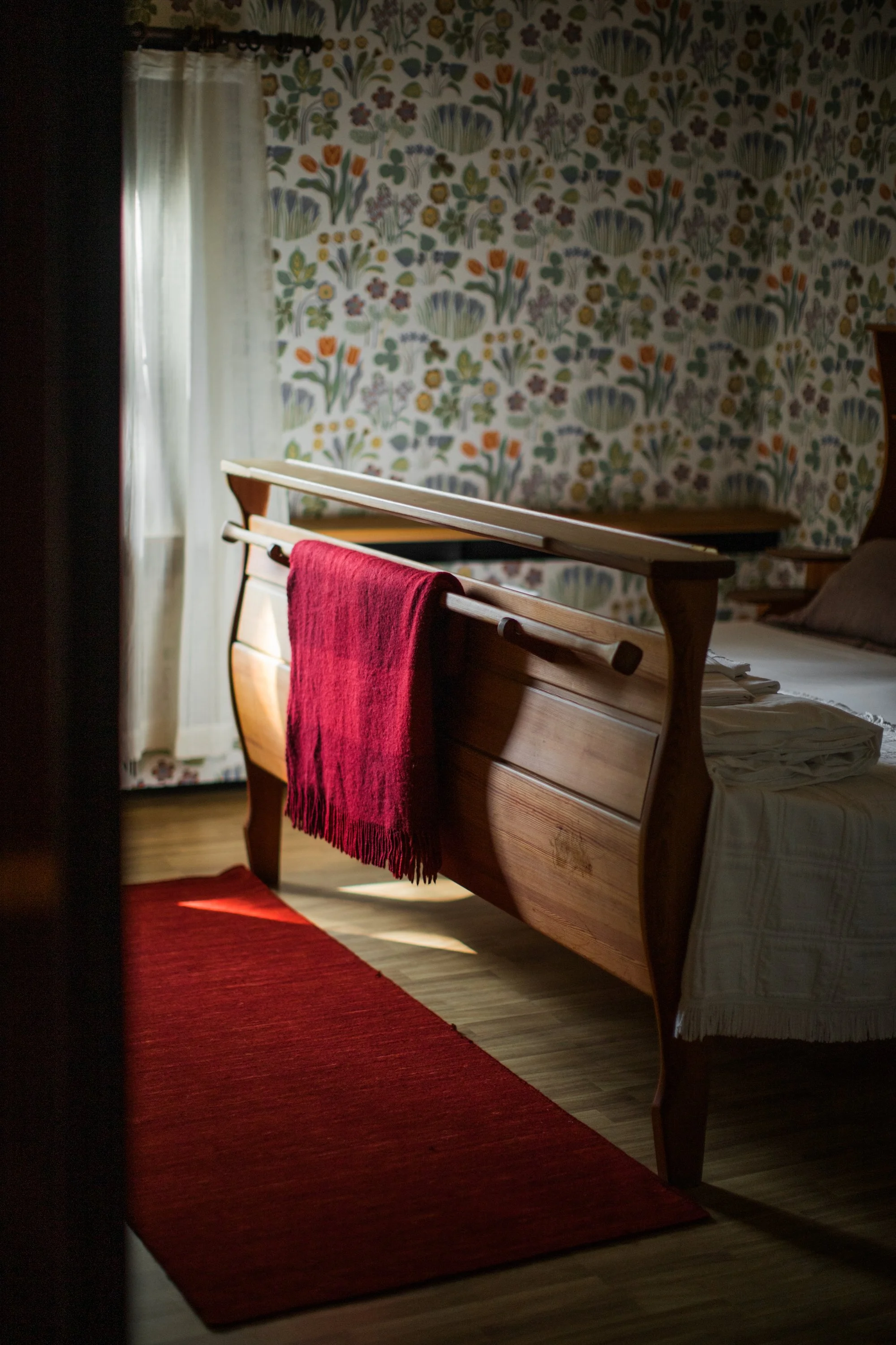 Ett sovrum med en trä säng, en röd filt, en matta och en tapet med blomsterallover mönster.