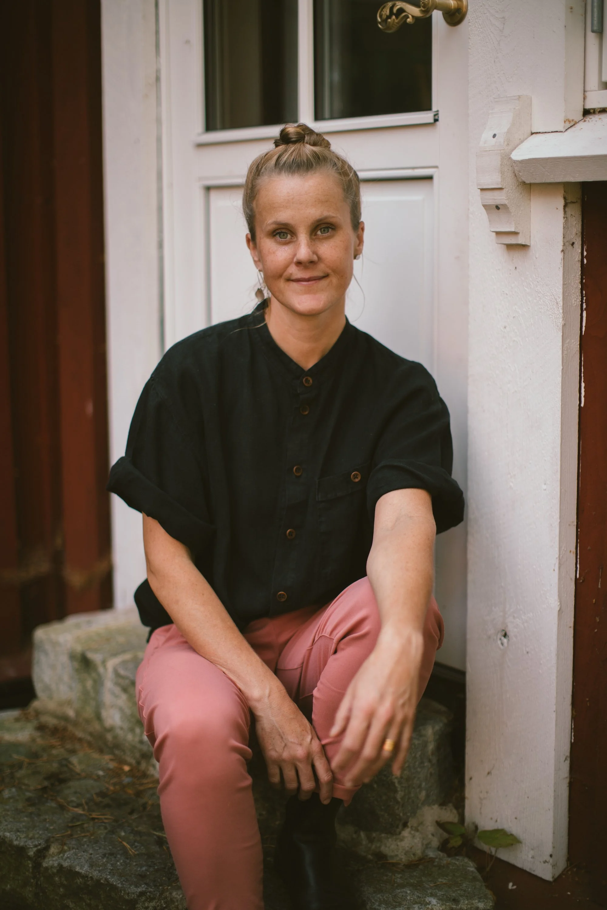 En kvinna sitter på en sten utanför en vit dörr och tittar mot kameran. Hon har hennes hår i en knut och bär en svart skjorta och rosa byxor.