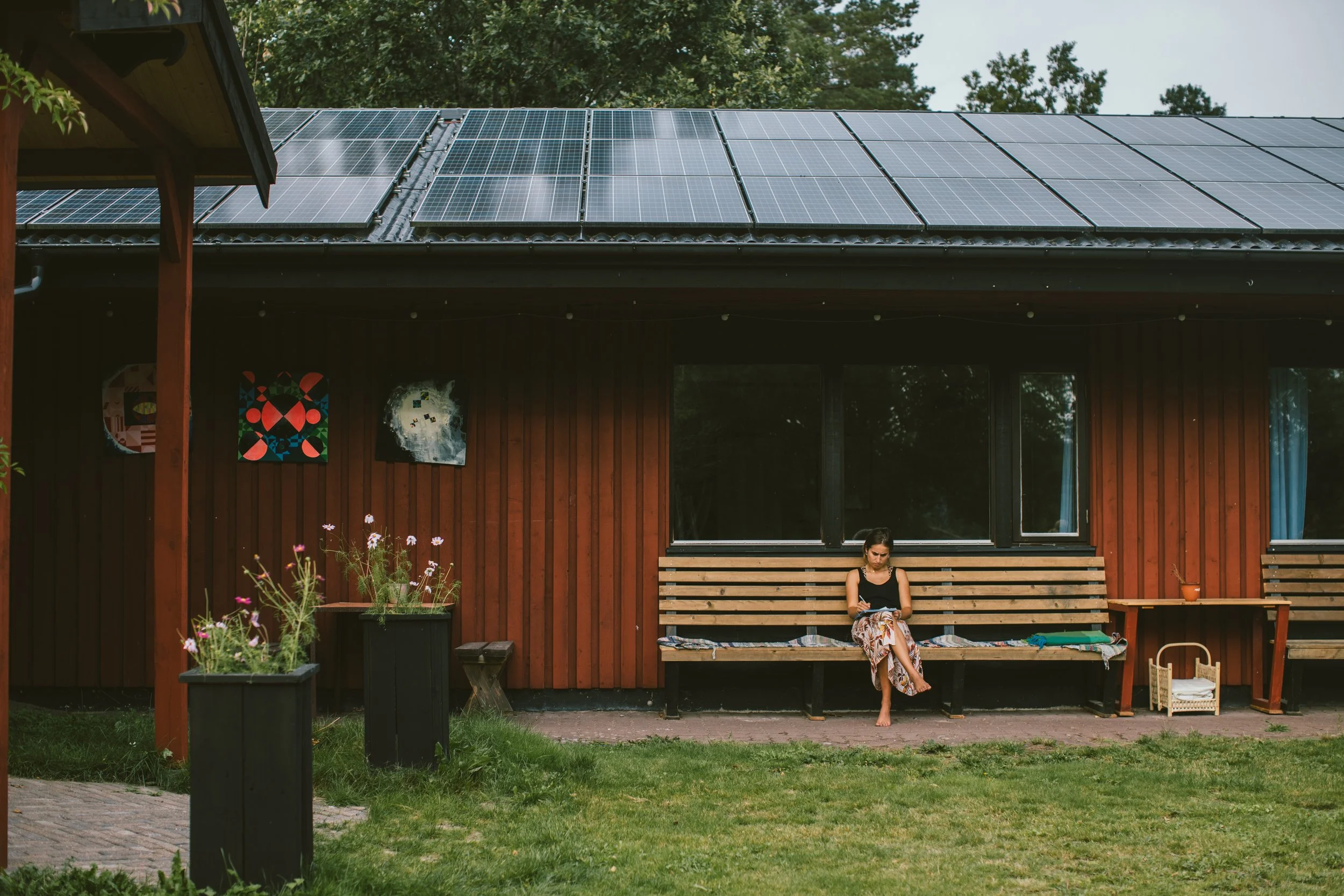 En person sitter på en träbänk utanför ett rött hus med solpaneler på taket. Det finns blommor i krukor framför och gröna träd i bakgrunden.