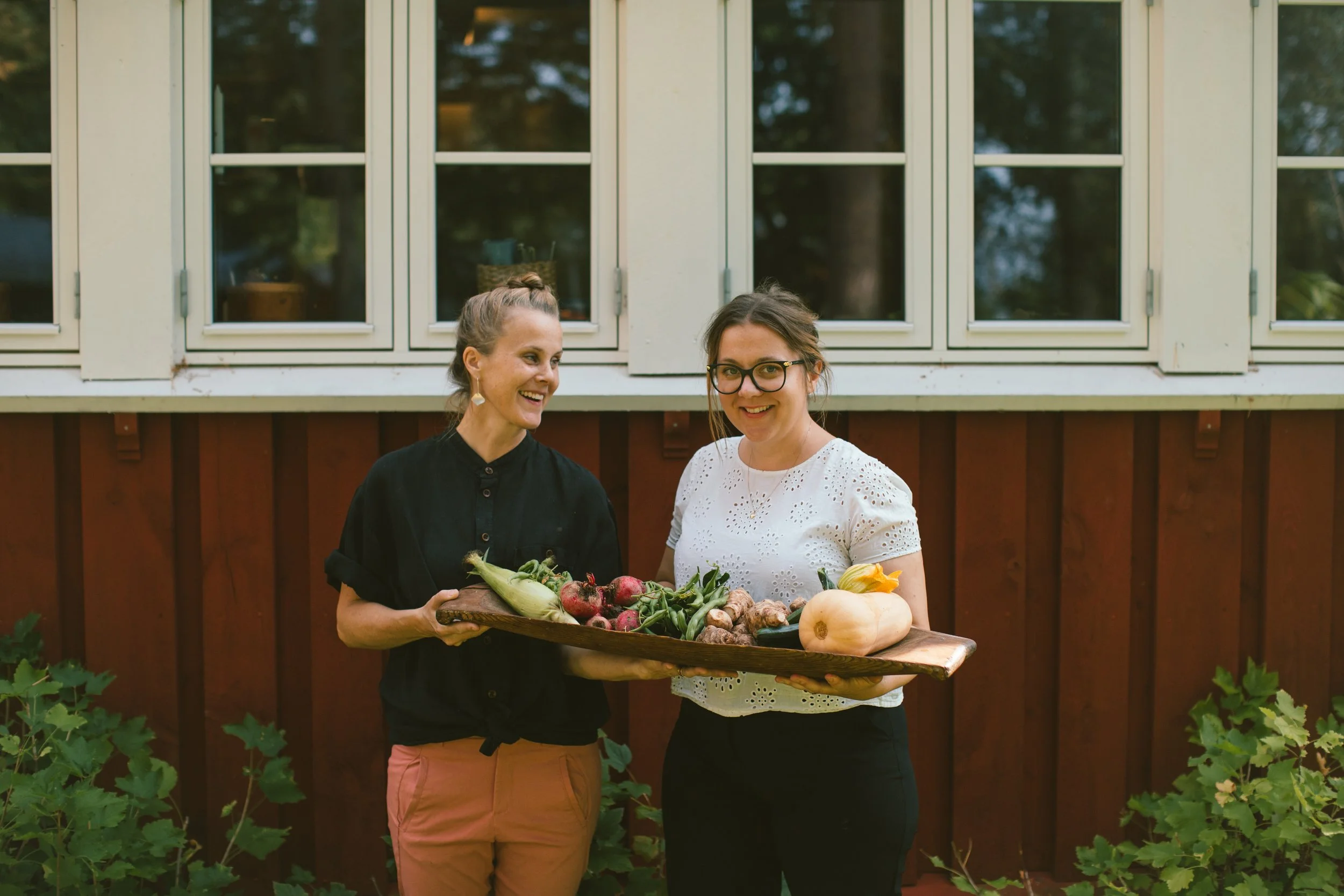 Moa och Mimi står utomhus framför ett rödbrunt hus med vita fönster, håller en stor träbricka med grönsaker som rädisor, kålrabbi och svamp, och ler.