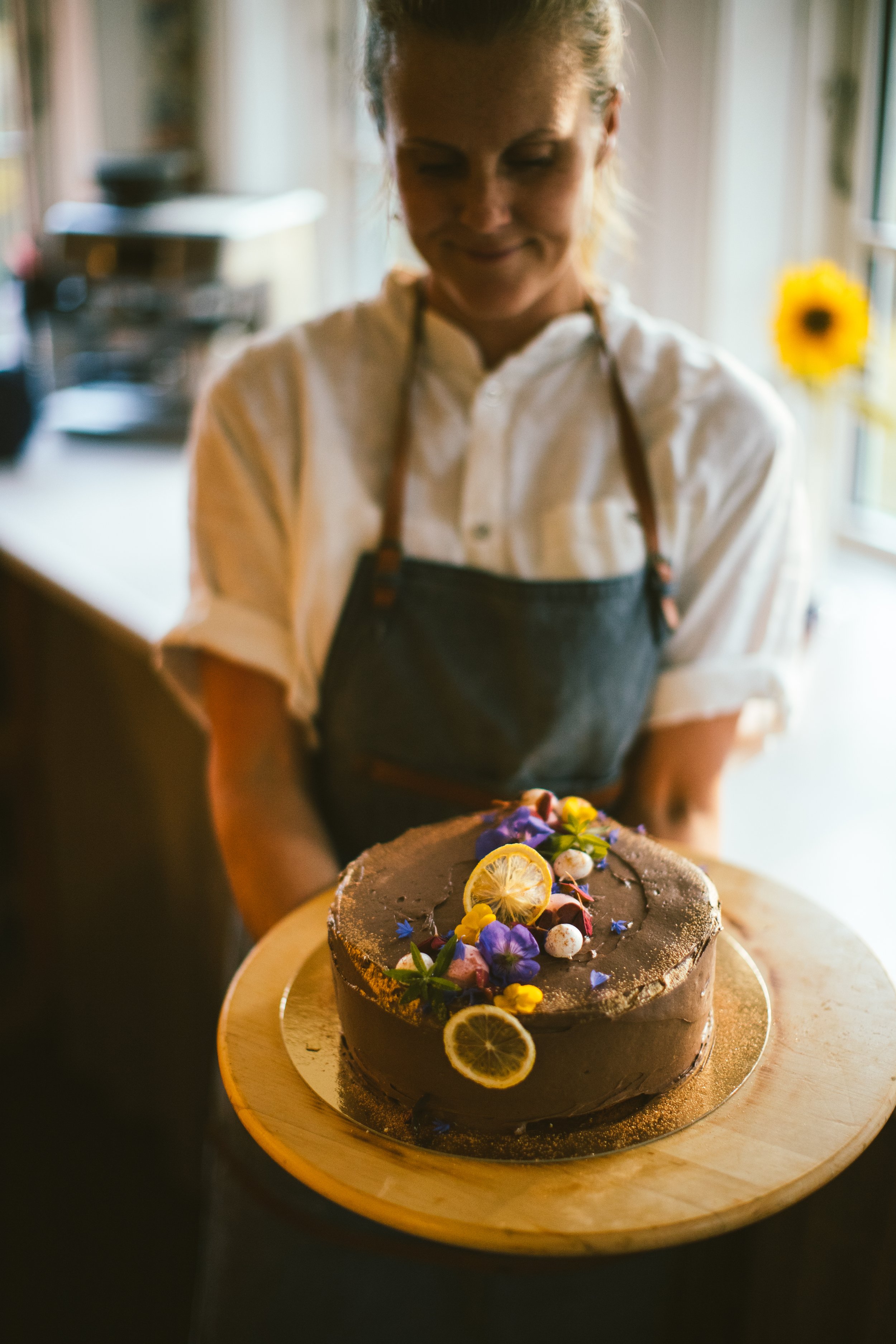 En kvinna i vit skjorta och svart förkläde som serverar en chokladtårta dekorerad med skivor av citron, blomster och små prydnadsföremål i ett kök.
