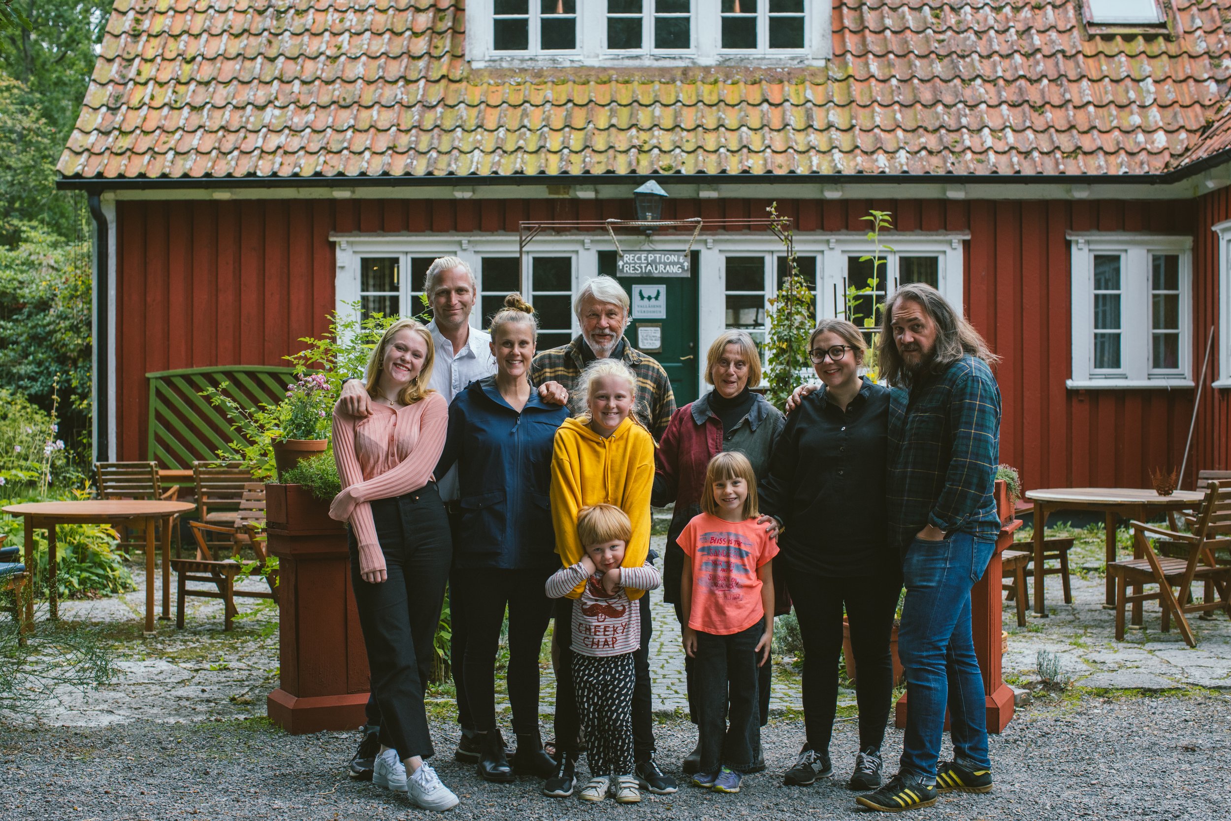 En grupp människor på familjeföretaget Vallåsens Värdshus står framför ett rödmålat hus med vita fönster, utomhus, omgiven av grönska.