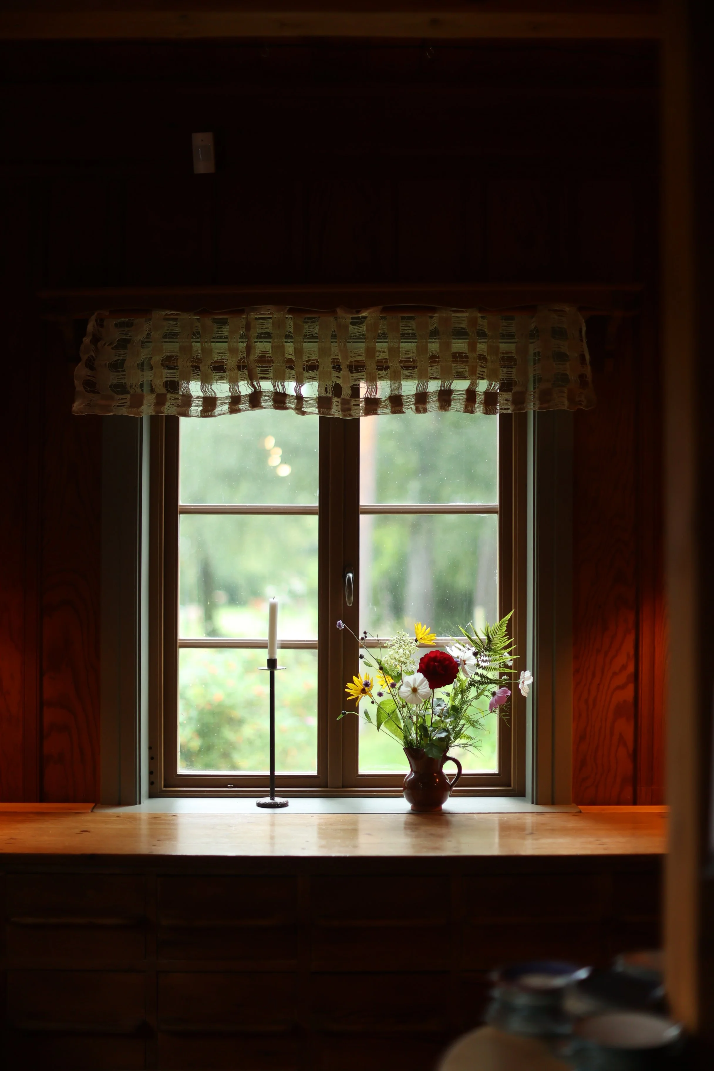 Fönster med gardin, blomsterarrangemang i vas, ljus, och utsikt över grön trädgård utomhus.