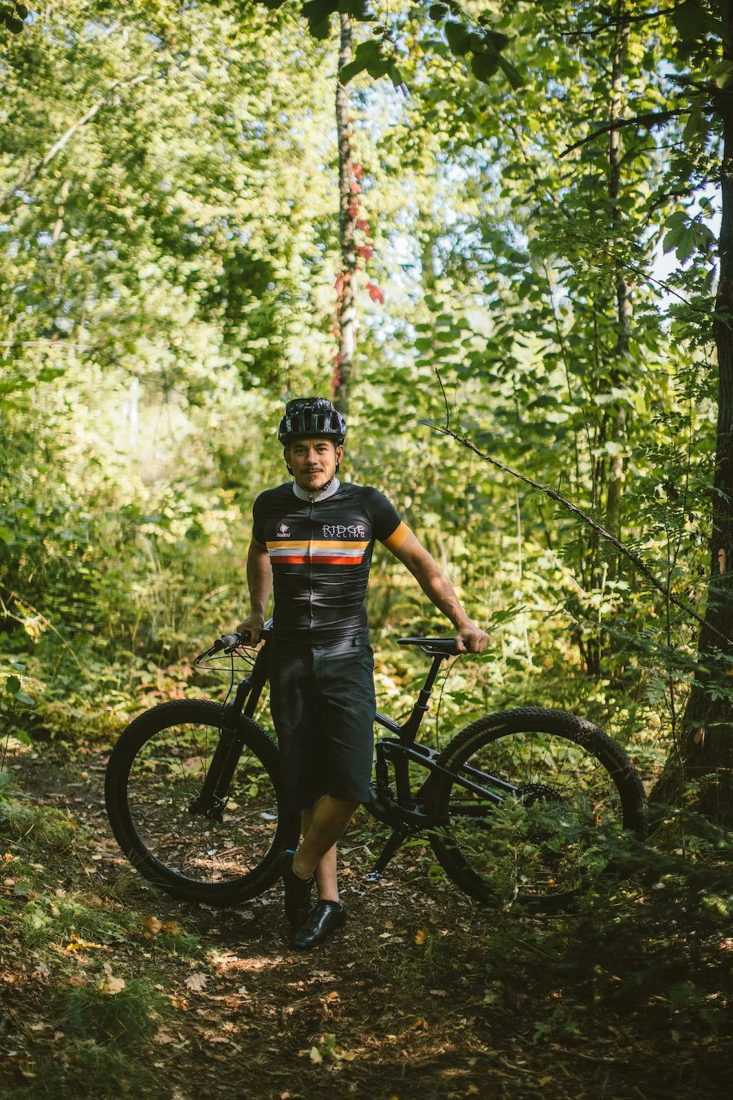 En man med mountainbike står i en skog, bär hjälm och sportkläder.