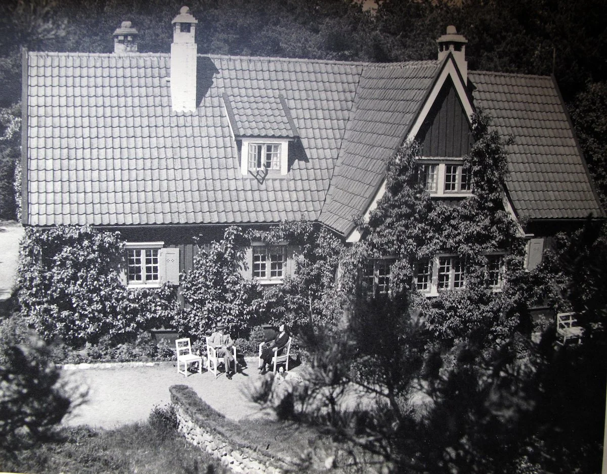 En svartvit bild av ett hus, Vallåsens Värdshus, med tak täckt av tegelpannor, omgiven av träd och buskar, med två personer som sitter på bänkar i trädgården.