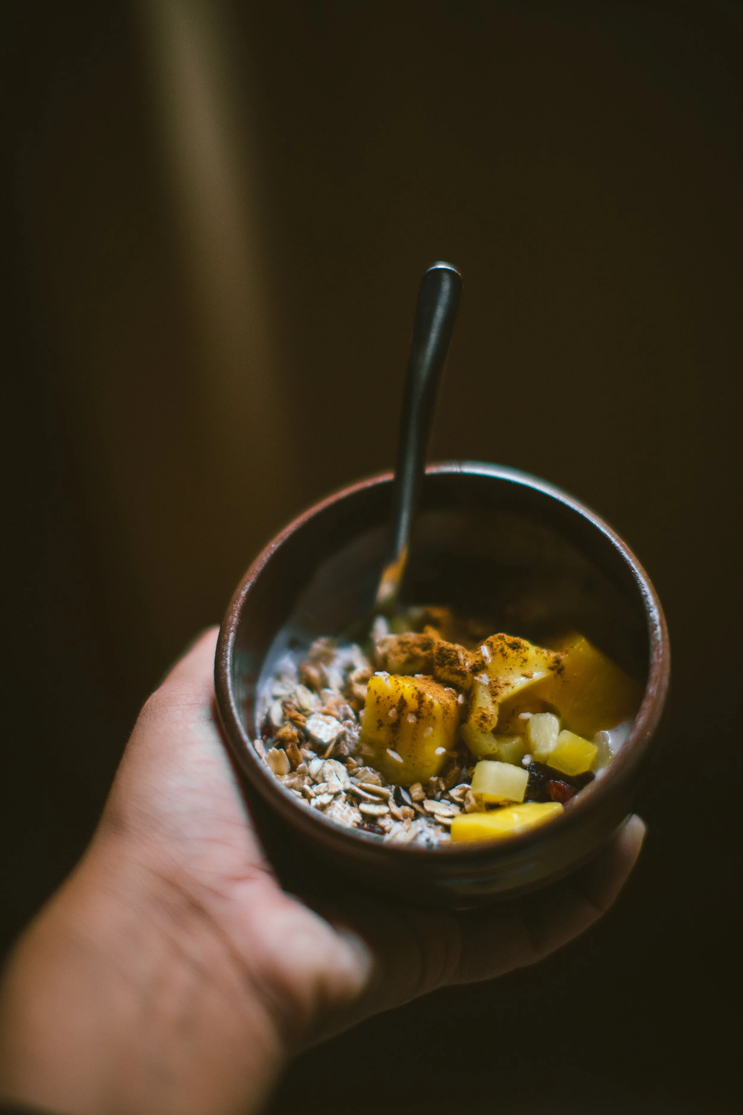 En hand håller en kopp fylld med havregryn, skivad banan och yoghurt, med en metallsked i koppen.