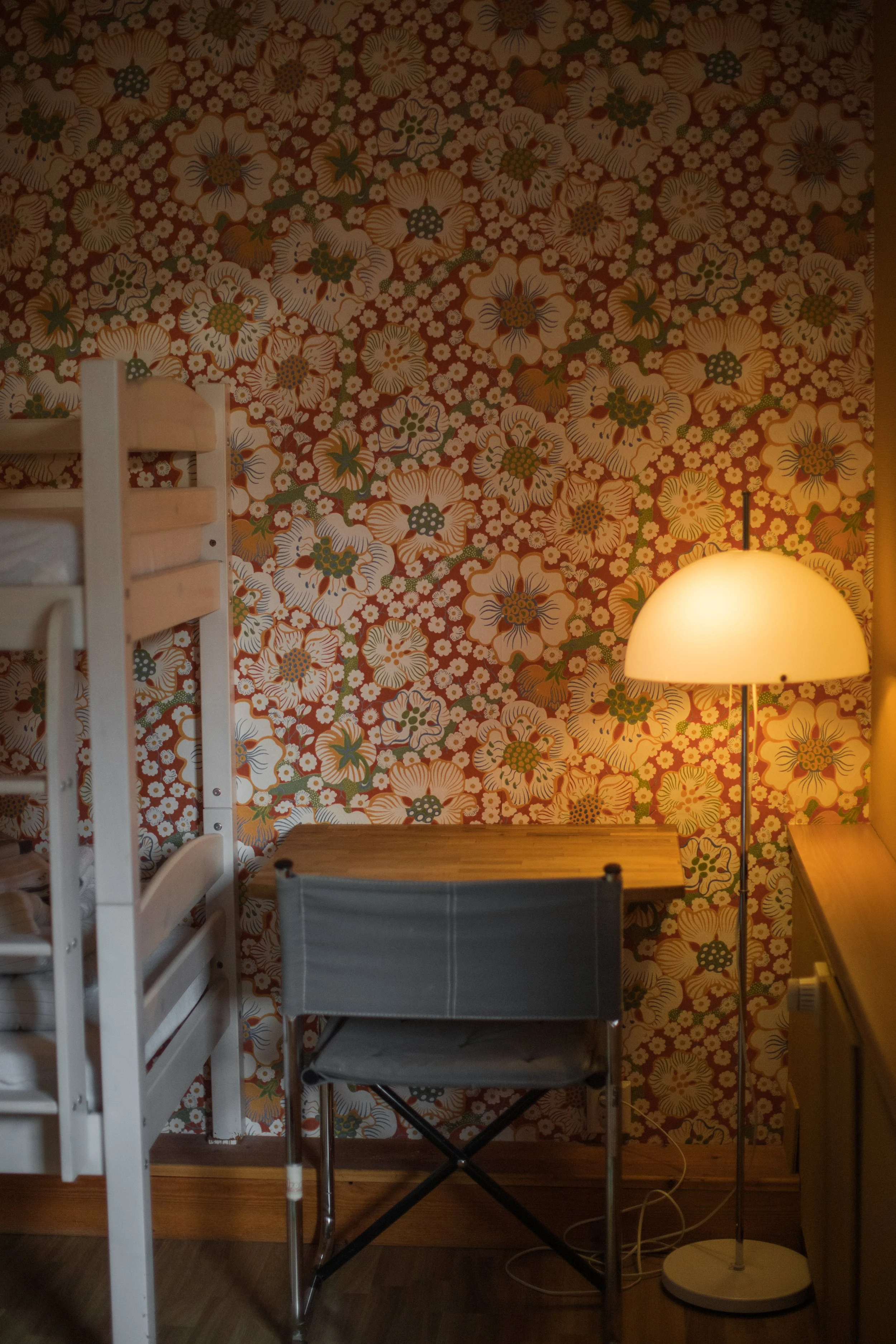 Ett rum med blommig tapet, en vit våningssäng till vänster, ett litet bord i mitten och en golvlampa till höger, som lyser.