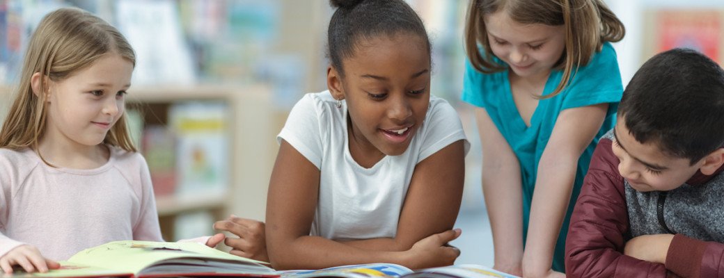 Four children, three girls and one boy, gathered around a book and reading together in a classroom setting.