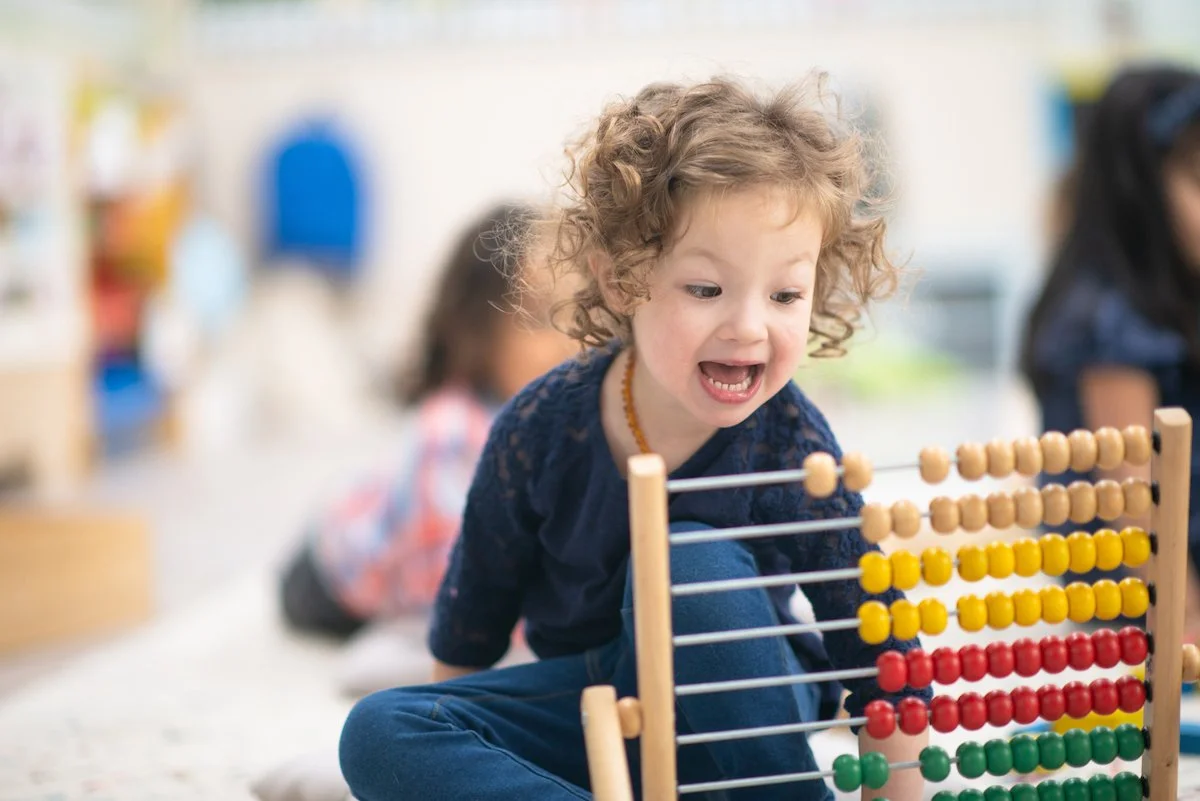 Child with curly hair playing with a colorful abacus in a classroom.