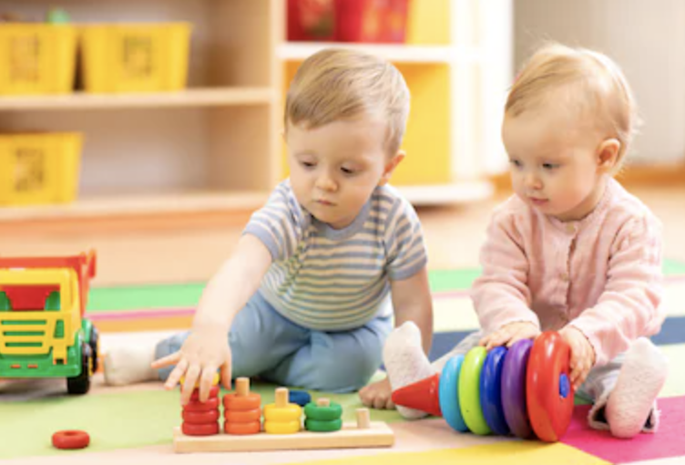 Two young children playing with colorful stacking toys on a multicolored mat in a playroom.