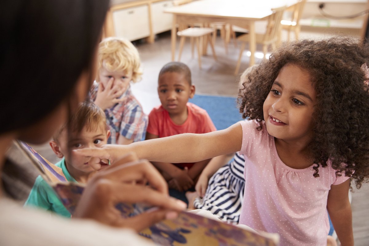 A young girl reaches out and touches an adult's hand while sitting on the floor with four other children in a classroom setting.