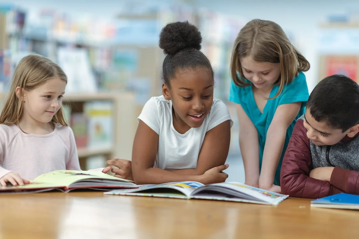 Four children are gathered around a table looking at colorful books in a library.