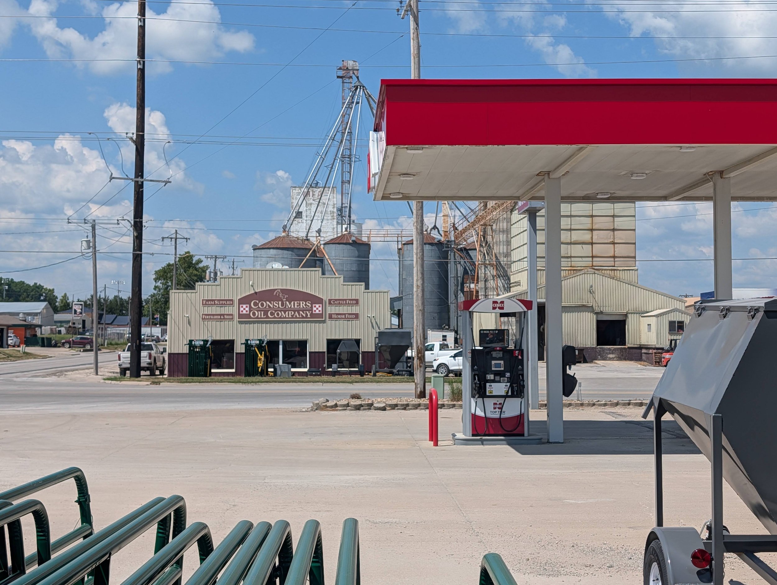 A gas station with a red roof, gas pumps, and an empty parking lot in front. In the background, there are industrial silos, electrical poles, and a partly cloudy sky.