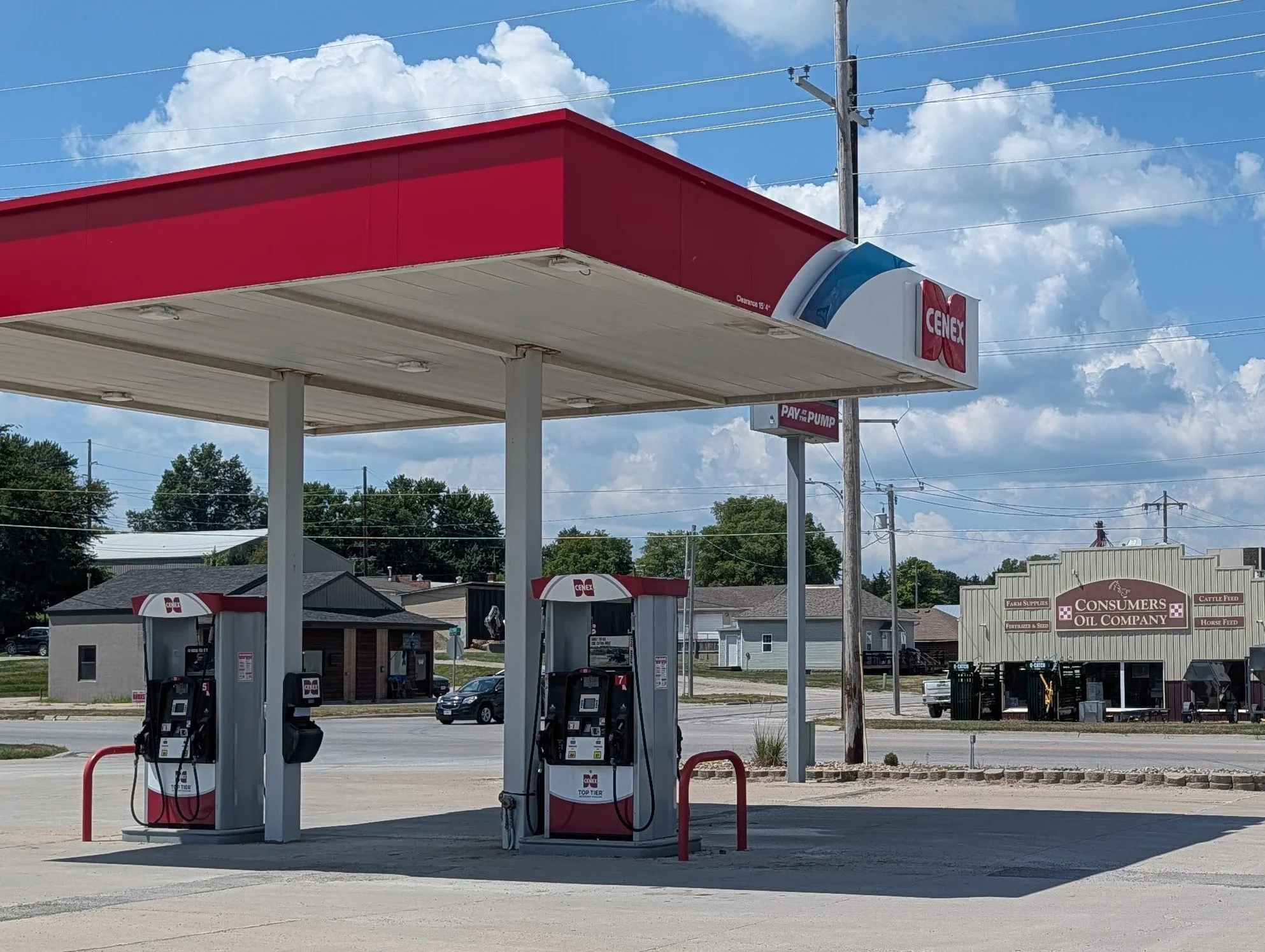 A gas station with multiple fuel pumps under a red and white canopy, with a small building and parking lot behind it, set against a background of trees and a partly cloudy sky.