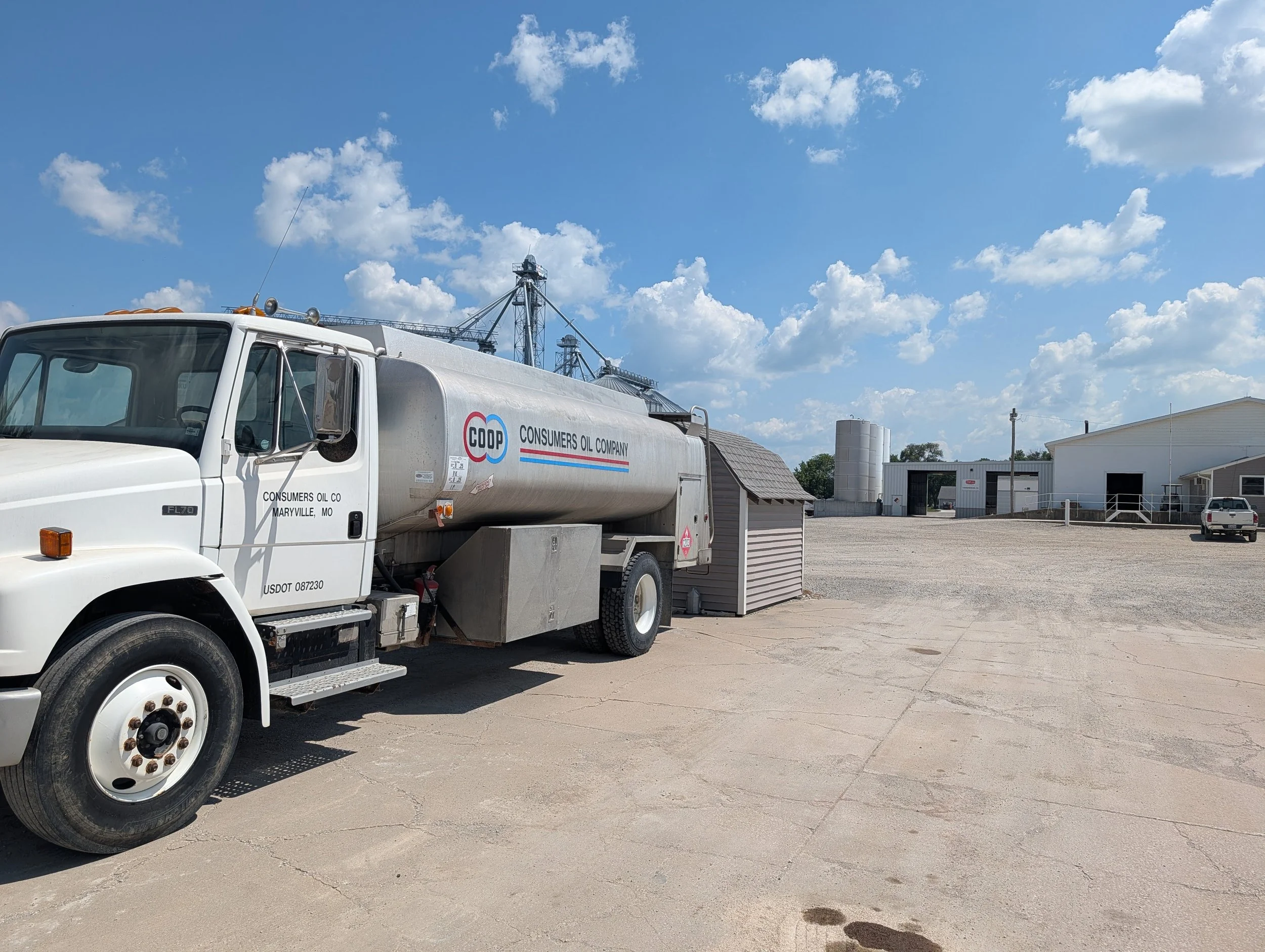 A white fuel delivery truck with 'Consumers Oil Company' and logo on the side parked on a concrete lot under a blue sky with scattered clouds, near a small building, a water tower, and farm structures.