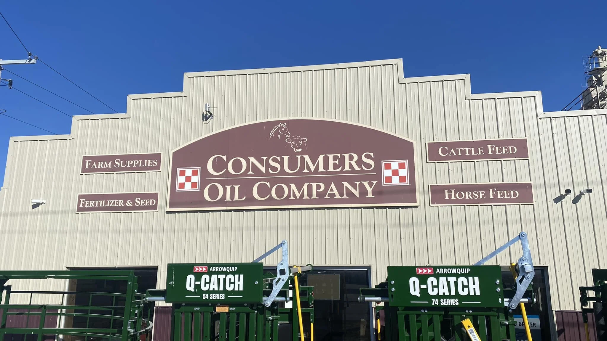 The exterior of a building with a sign for Consumers Oil Company, offering farm supplies, cattle feed, fertilizer and seed, and horse feed, with blue skies above.