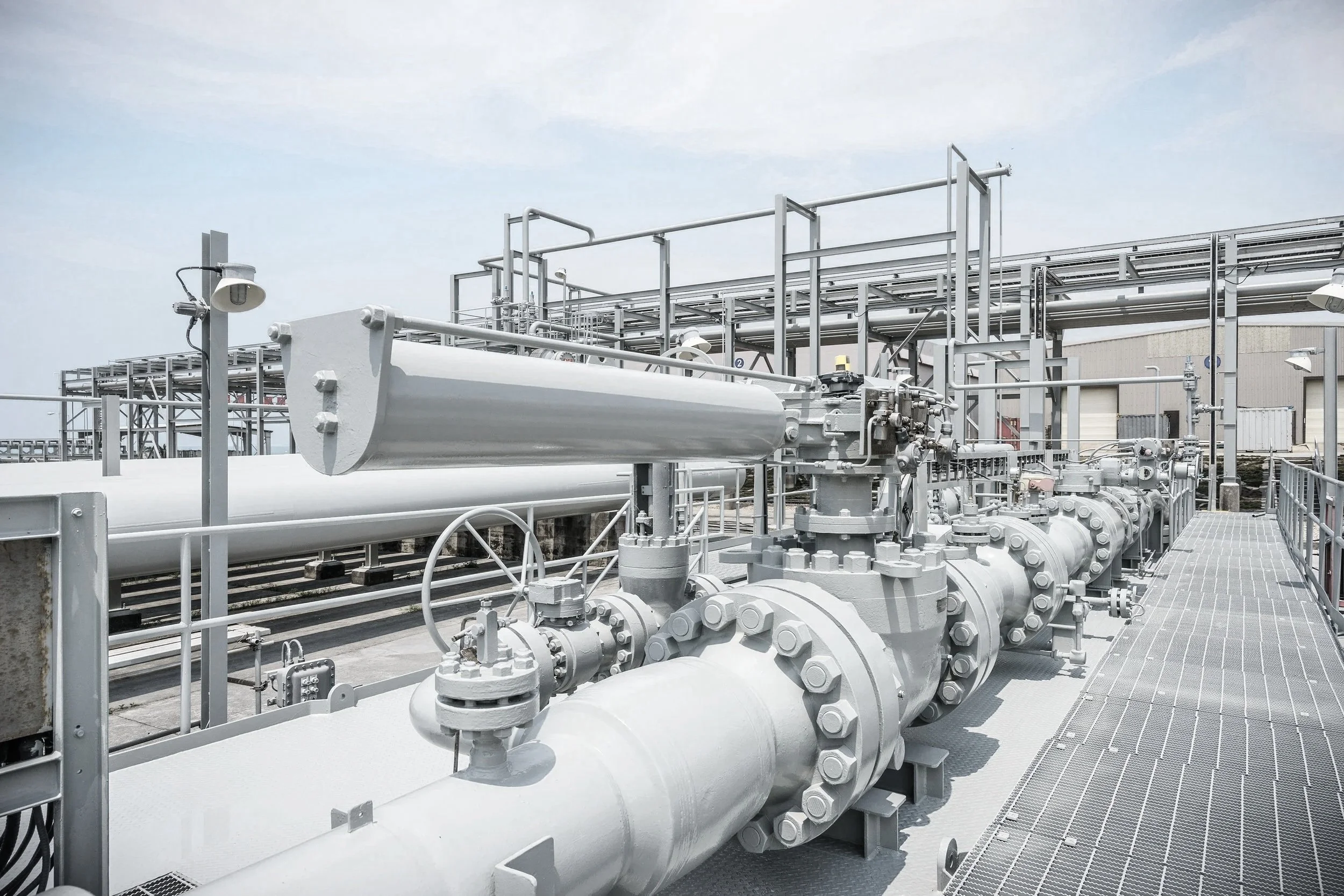 Industrial facility with large gray pipes, valves, and metal framework under a partly cloudy sky.
