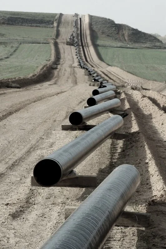 Large black pipes laid out in a row along a dirt road leading up a hill, with construction equipment at the top, under a gray sky.