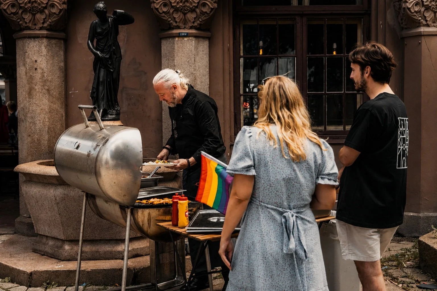 En gruppe mennesker står foran en grill med mat, en person tilbereder mat, mens to andre venter, en person har et pride-flagg, og de er utenfor en bygning med en statue og vindu i bakgrunnen.