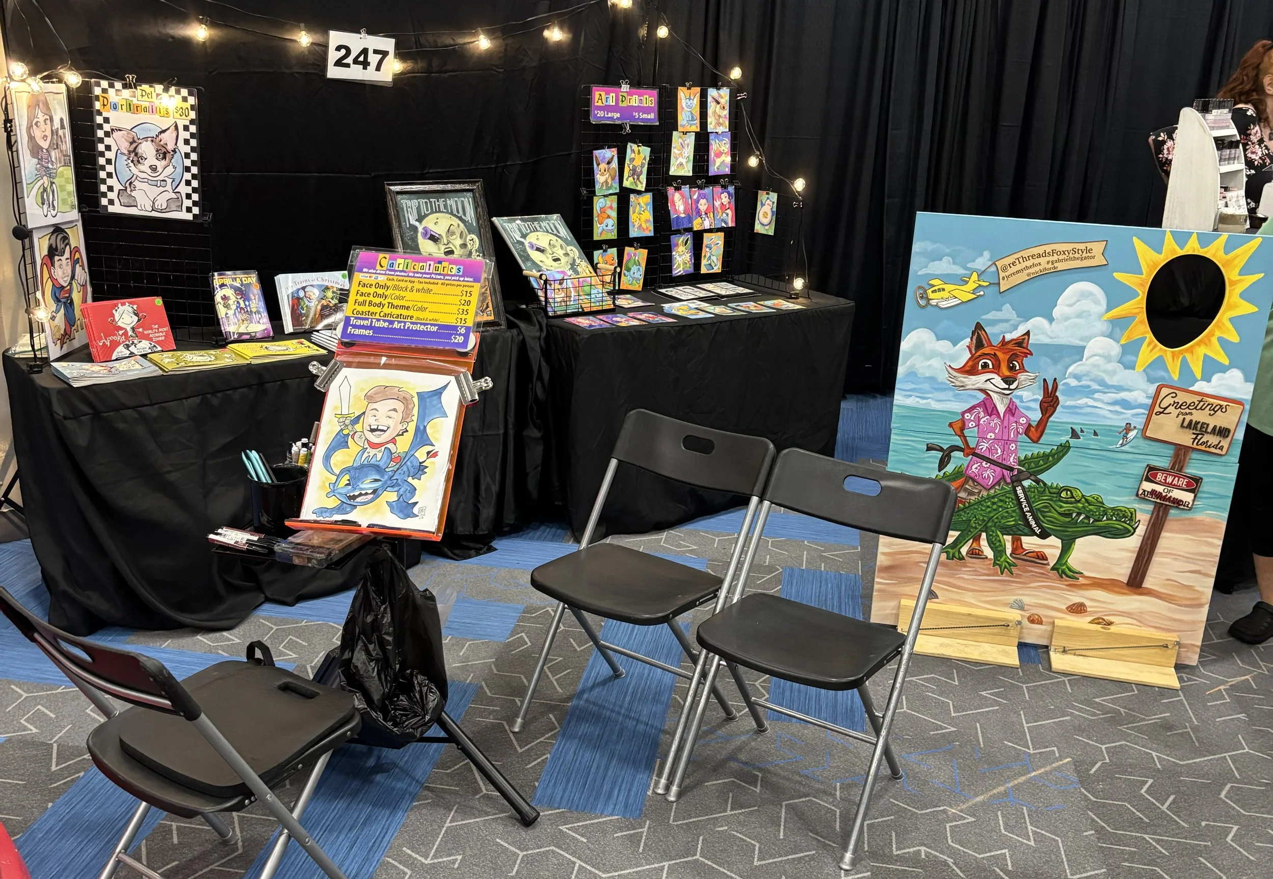 Booth at an art fair with colorful artwork and crafts, including paintings of fantasy characters, animal illustrations, and a large painted sign of a fox in a tropical scene. The booth has string lights and price signs, with some chairs in front.