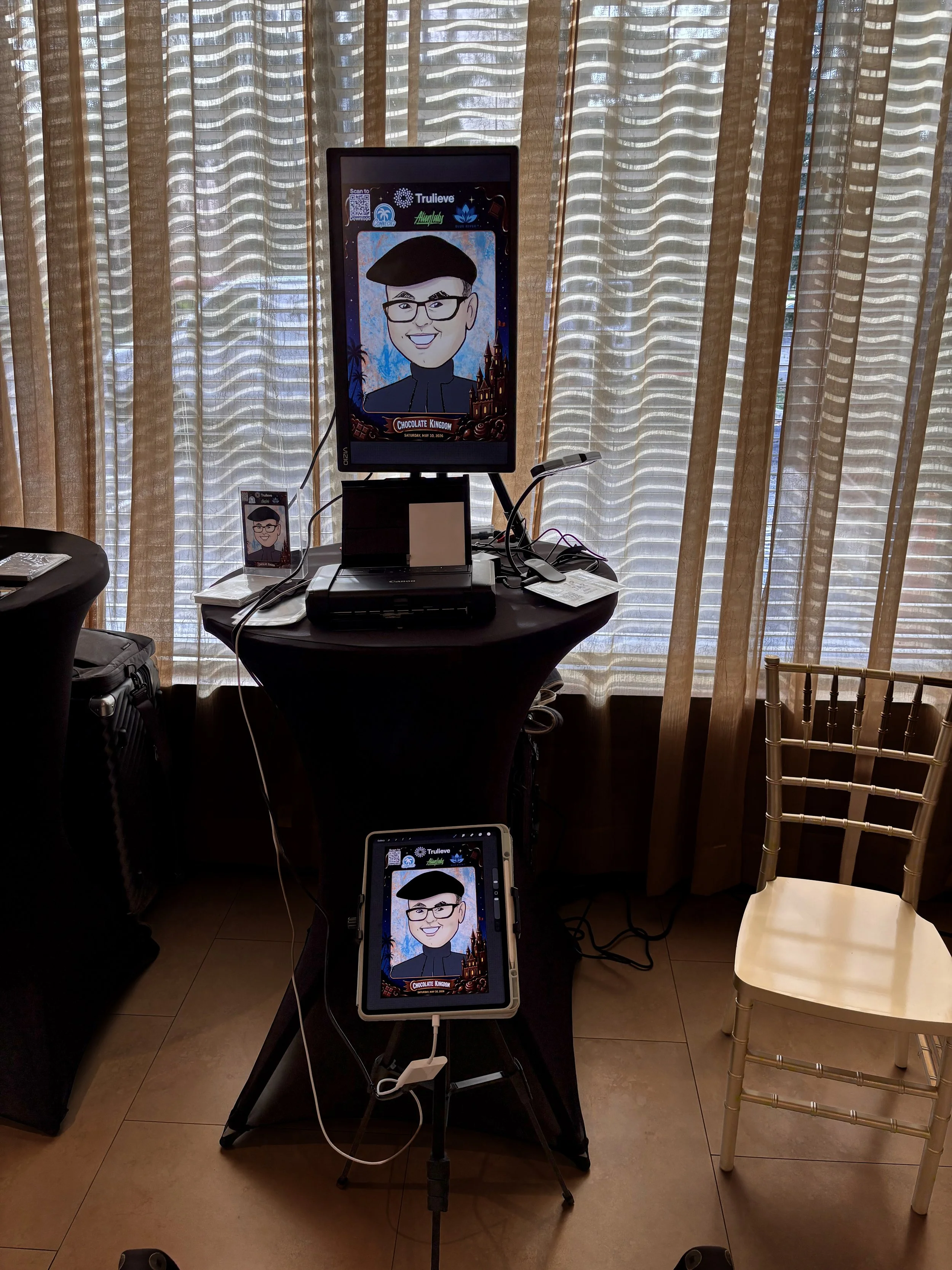 A digital cartoon portrait of a man with glasses and a hoodie displayed on a large screen, with a similar cartoon image on a smaller phone screen placed on a black table. The background has beige curtains with a wavy pattern, and surrounding objects 