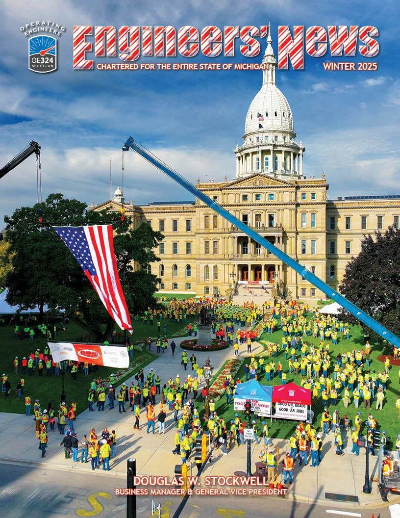People wearing yellow vests gather outside the Michigan state capitol building. Two cranes are lifting an American flag and another banner, with a crowd holding signs and banners in the foreground.