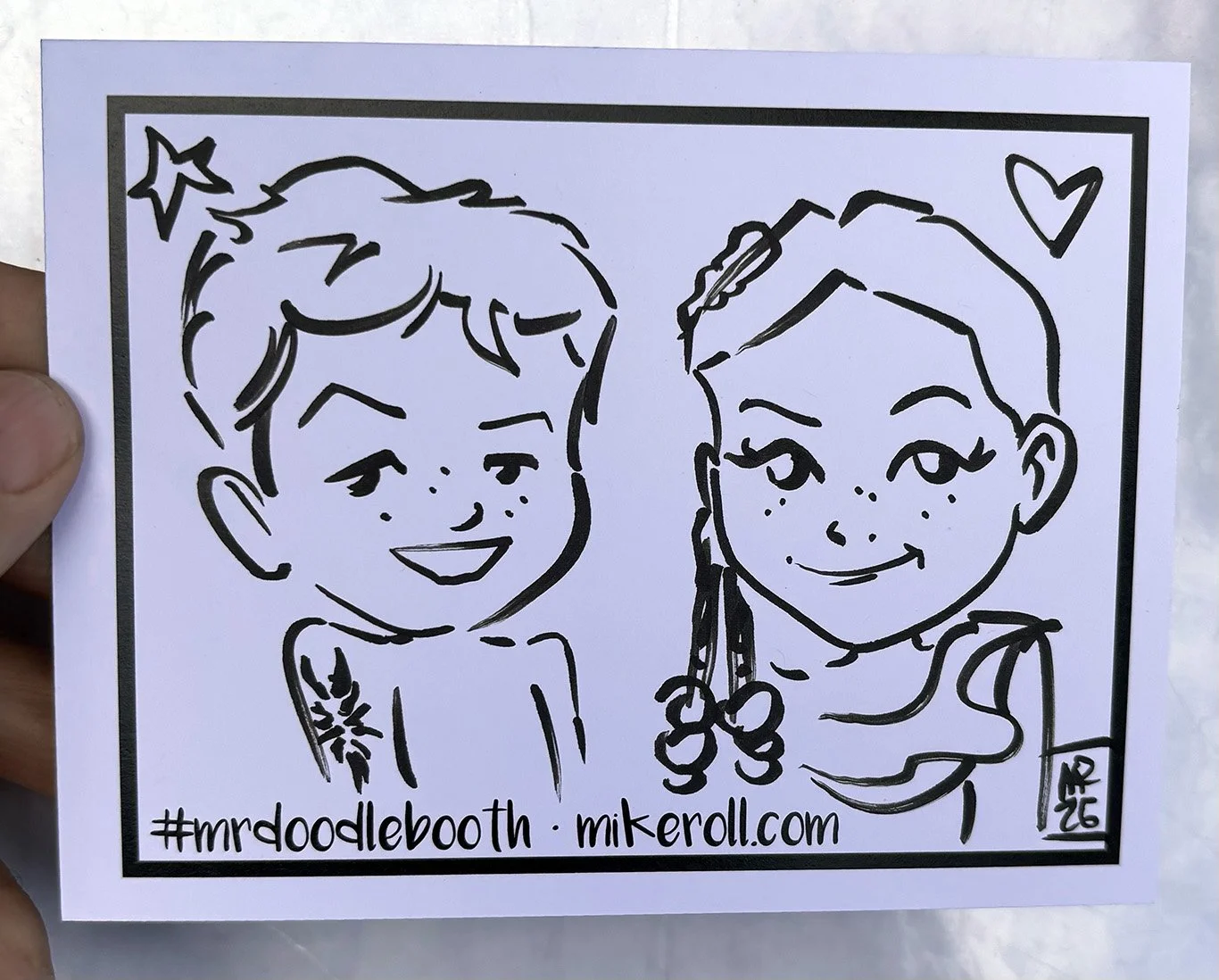 A black and white line drawing of two smiling children, a boy on the left with short hair and a girl on the right with long hair and cherries in her hair. There is a small heart in the top right corner and a star in the top left corner. The drawing i