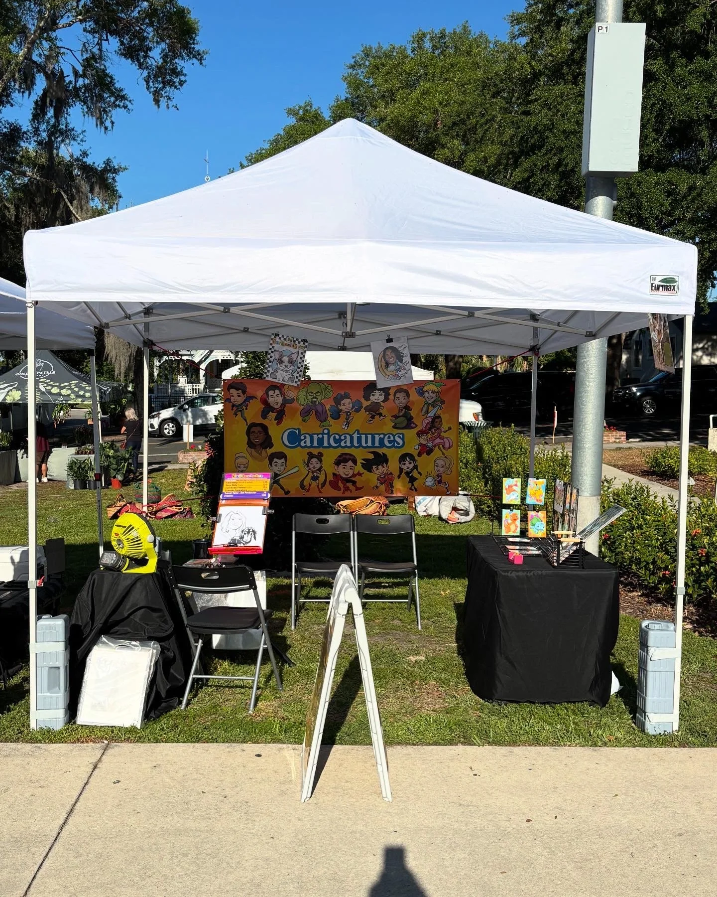 Outdoor art booth with caricature illustrations displayed, tables with art supplies, chairs, and display items under a white canopy tent at a sunny park event.