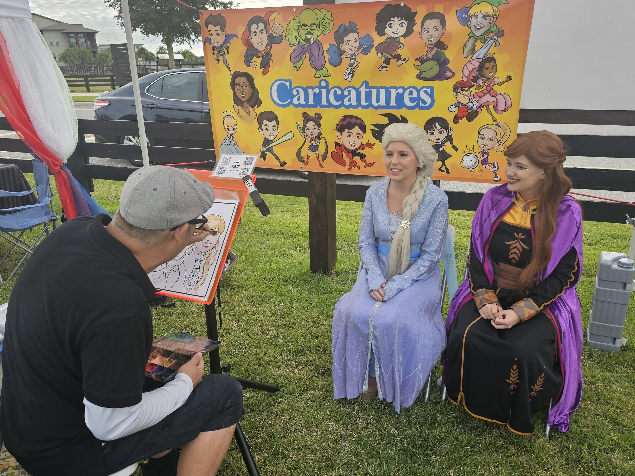 A caricature artist sketches a portrait of a woman dressed as a princess with two buns in her hair, sitting on a chair while two women dressed as characters from movies or fairy tales sit nearby. A colorful banner with cartoon caricatures of various superheroes, princesses, and fantasy characters is displayed behind them, with the word 'Caricatures' written prominently.
