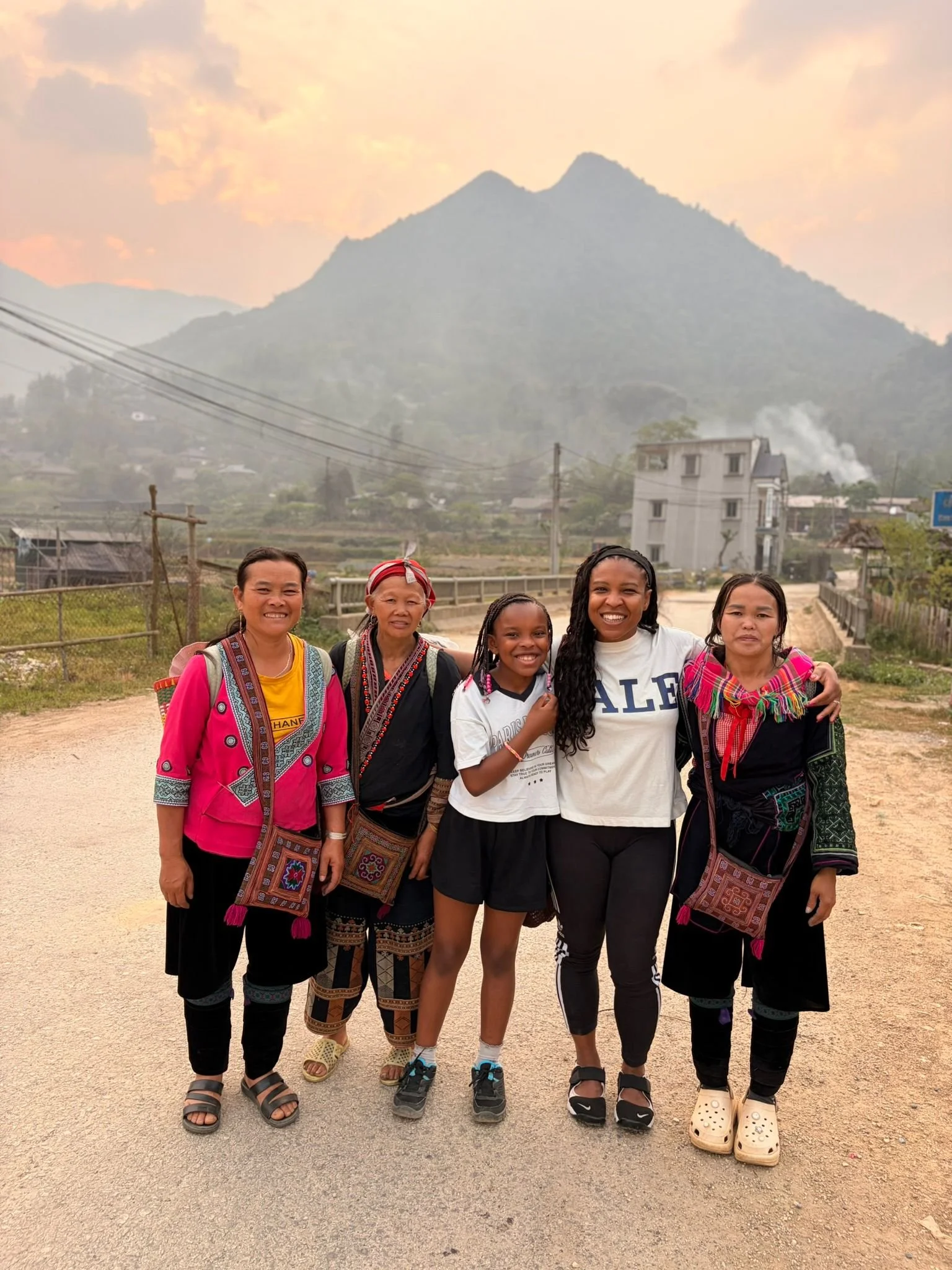 Groupe de cinq femmes et une jeune fille posant devant une montagne, en tenue traditionnelle colorée, dans un village rural.