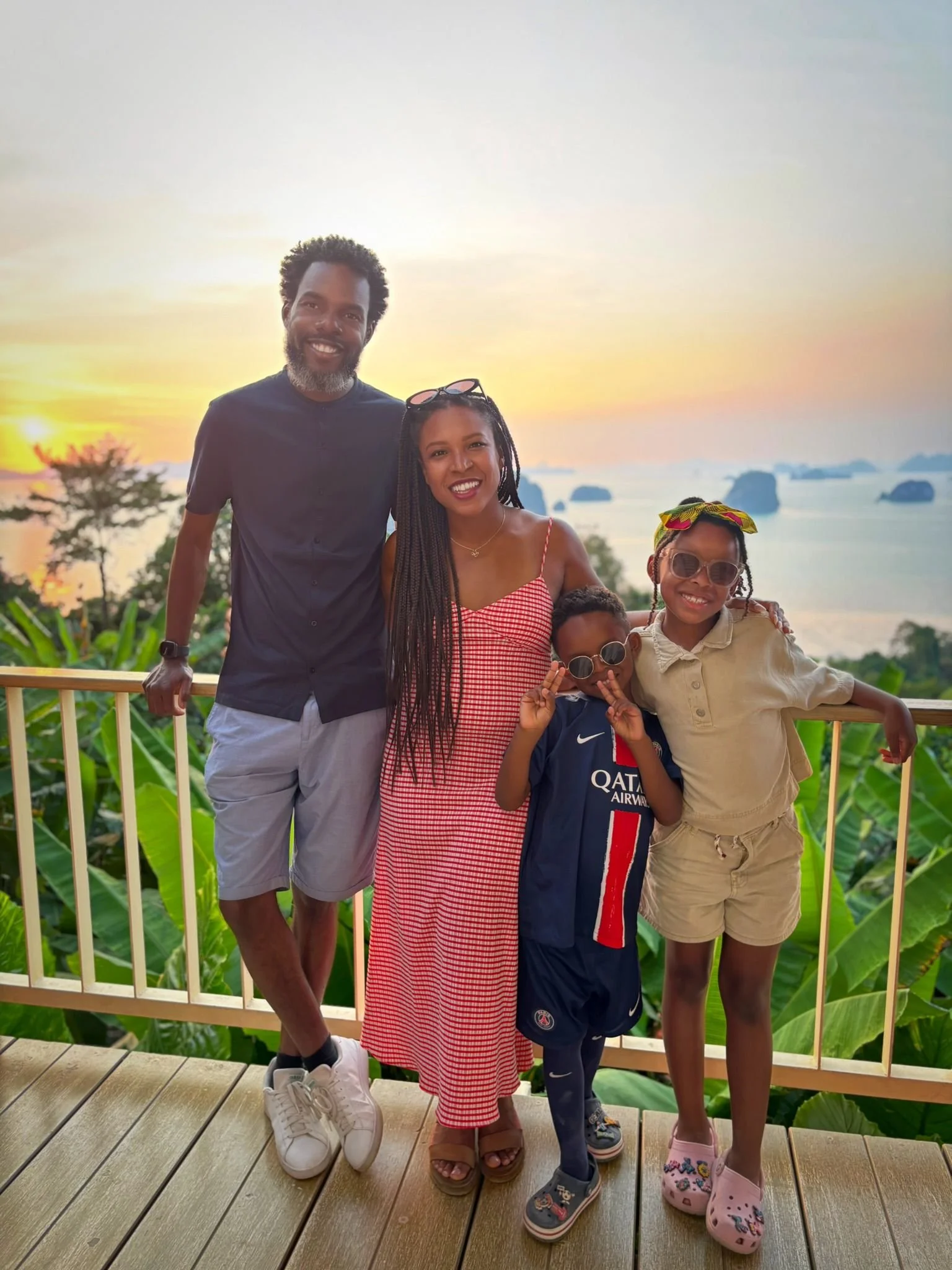 Un groupe de cinq personnes posant sur une terrasse en bois, avec un paysage de mer et d'îles au coucher du soleil en arrière-plan. Deux adultes et trois enfants souriants, portant des vêtements décontractés et des accessoires comme des lunettes de soleil.