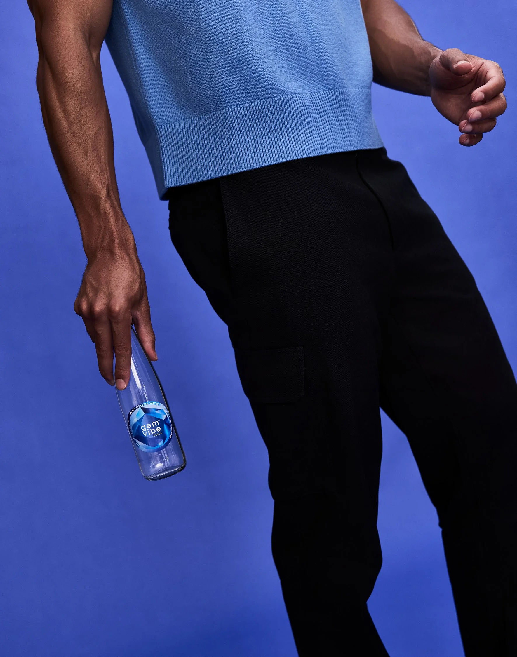 Nahaufnahme eines Mannes, der eine Flasche Mineralwasser hält, während er eine dunkle Hose trägt und vor einem blauen Hintergrund steht.