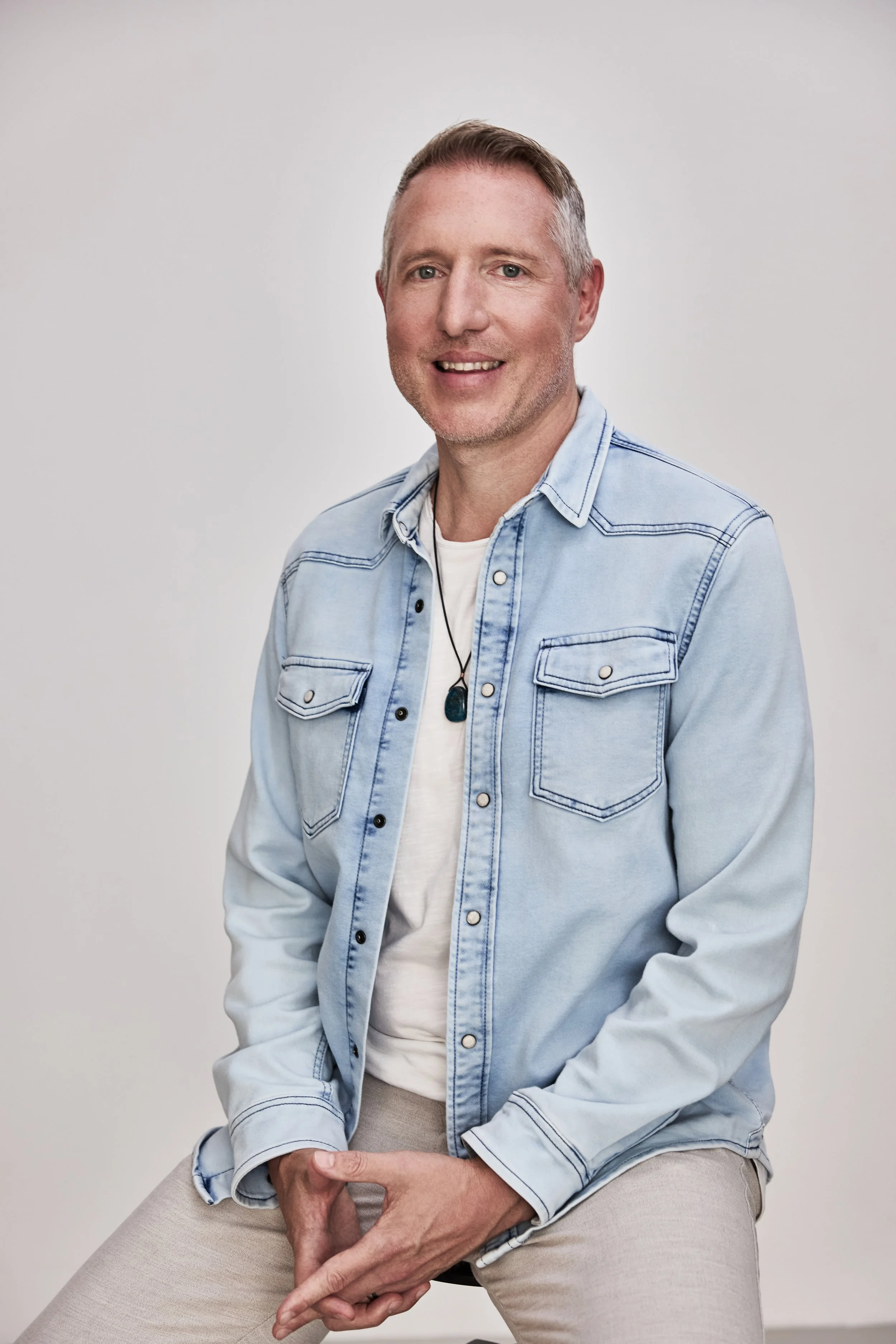 Ein Mann sitzt auf einem Stuhl, trägt eine hellblaue Jeansjacke und hellgraue Stoffhose, hat kurze braune Haare und lächelt. Background ist weiß.